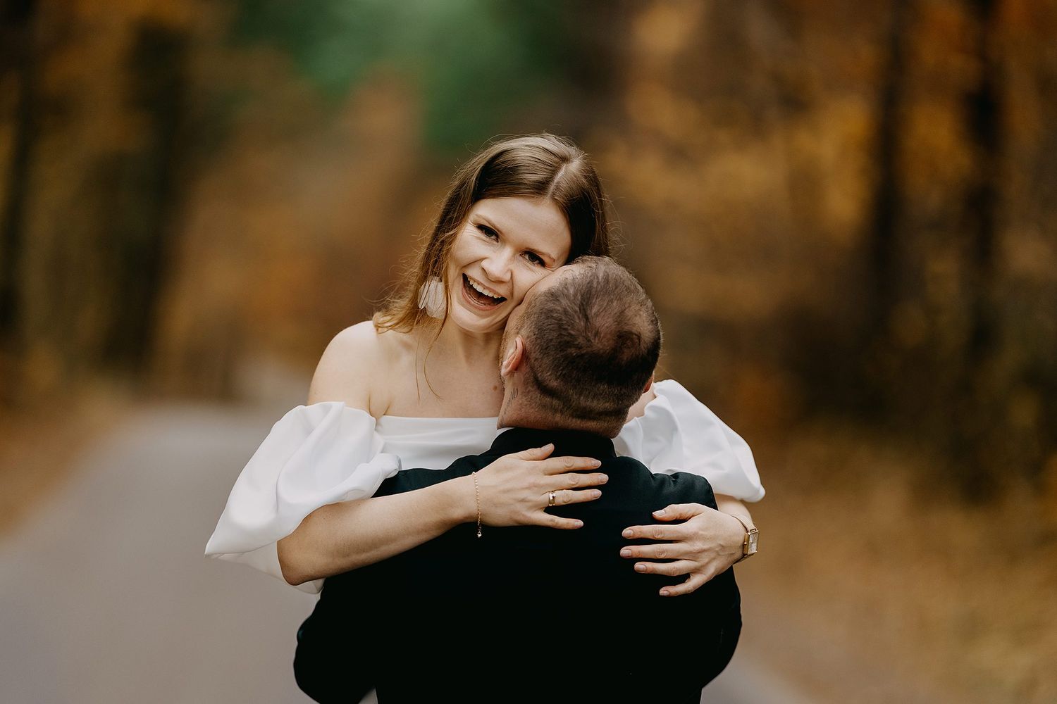 Joyful bride in white off-shoulder dress laughs while embracing groom on autumn road.