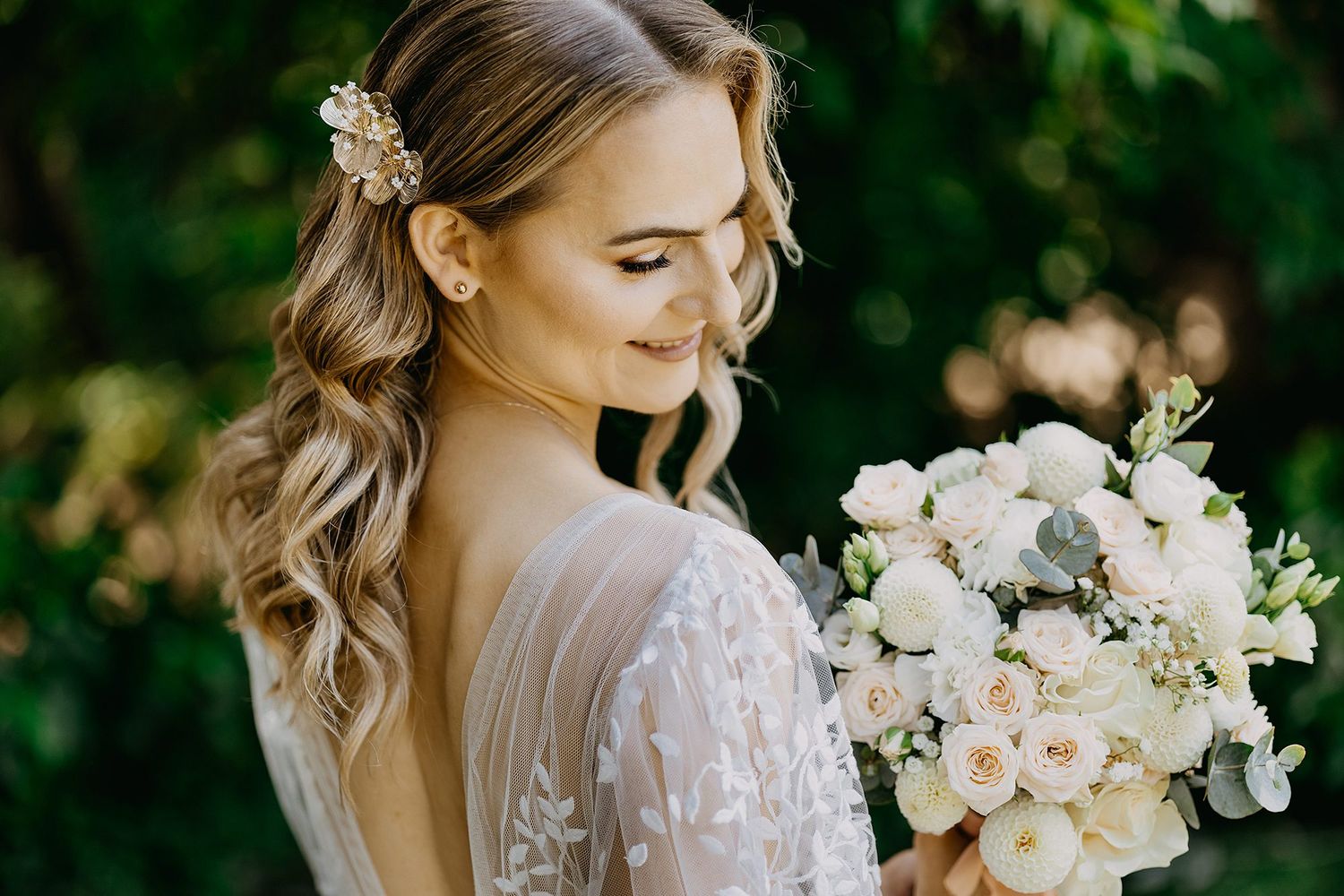 Elegant bride with wavy hair holding a white rose bouquet in a lace wedding gown outdoors.