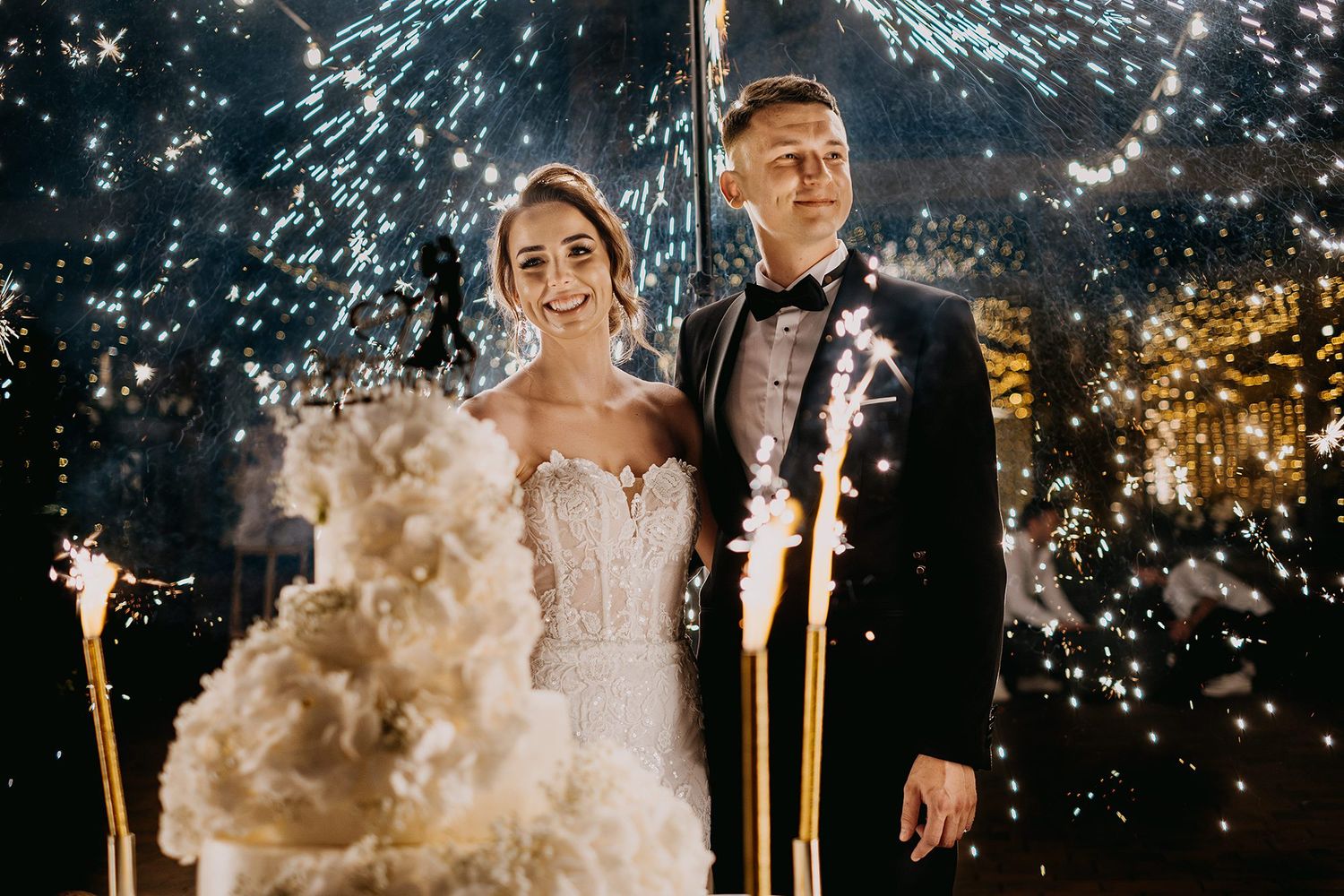 Bride and groom posing beside floral wedding cake surrounded by sparkling fireworks at night.