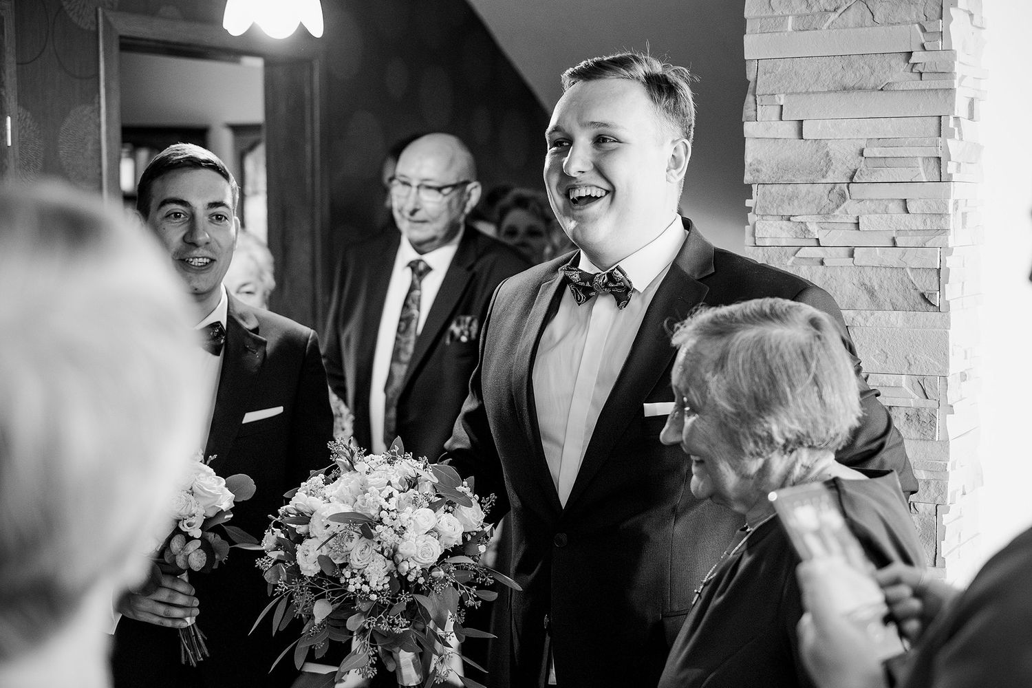 Black and white photo of smiling groom greeting guests while holding bridal bouquet at wedding.