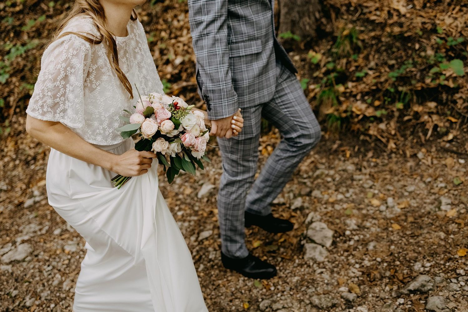 Bride in lace gown holds pink bouquet while walking with groom in plaid suit on rocky path.