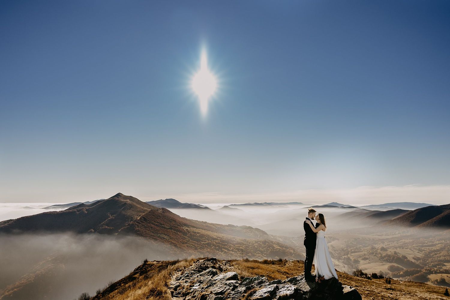 Wedding couple standing on mountain peak above clouds under bright sun.
