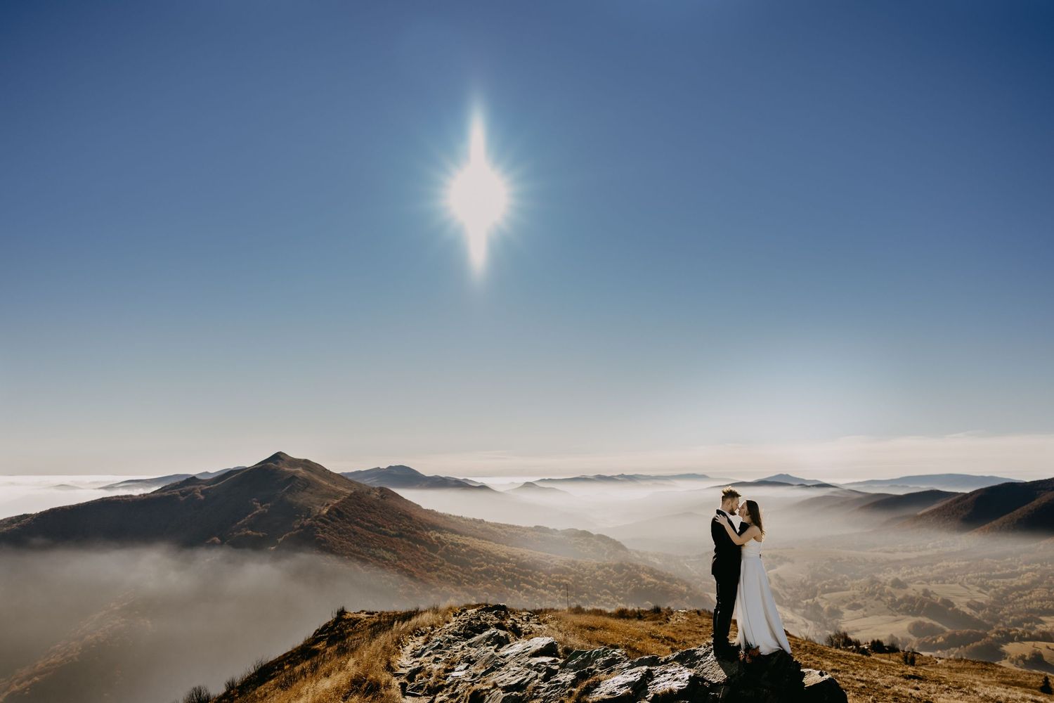 Bride and groom embrace on rocky mountain summit above clouds with bright sun star burst in blue sky.