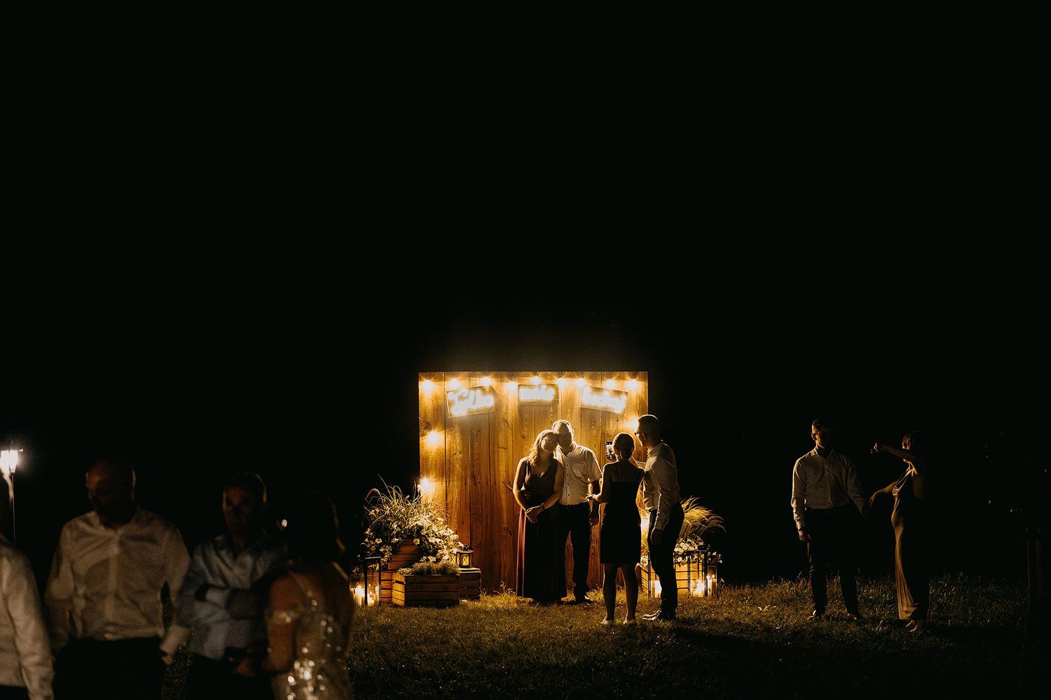Guests gather around a warmly lit wooden structure at an outdoor nighttime event or wedding reception.
