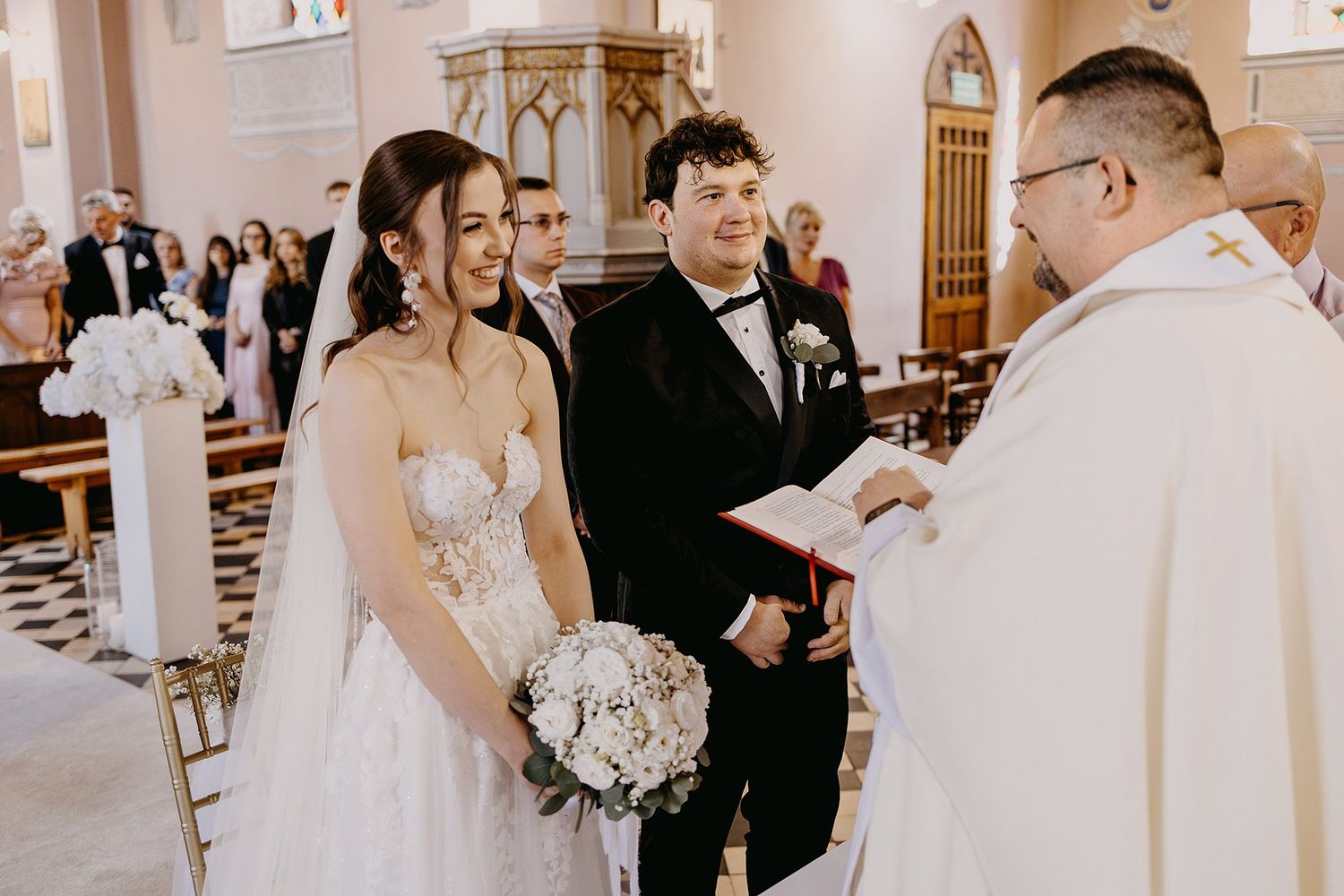 Smiling bride and groom exchange vows before a priest in a traditional church ceremony.