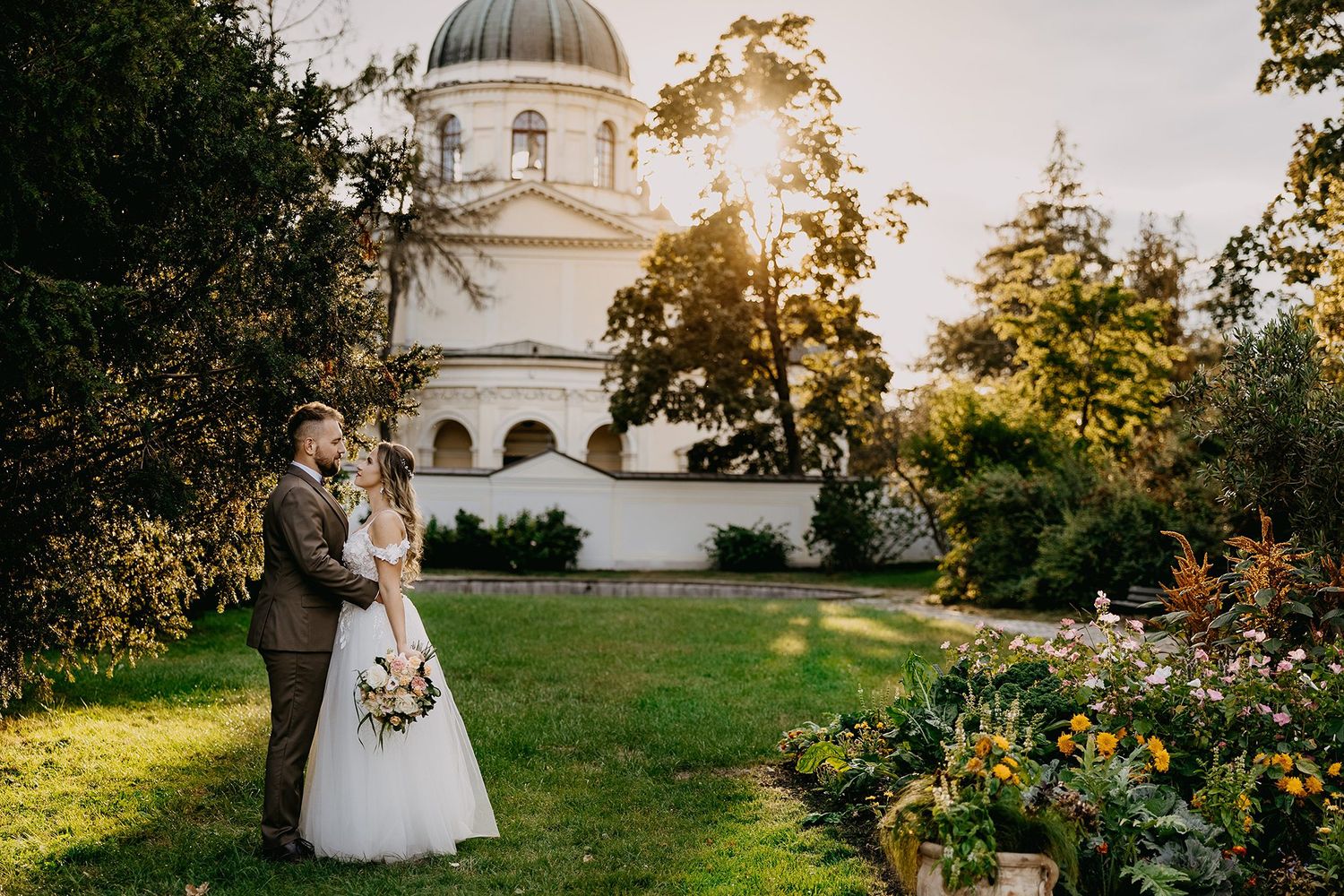 Wedding couple standing in a lush garden with a historic domed chapel glowing in golden sunset light.