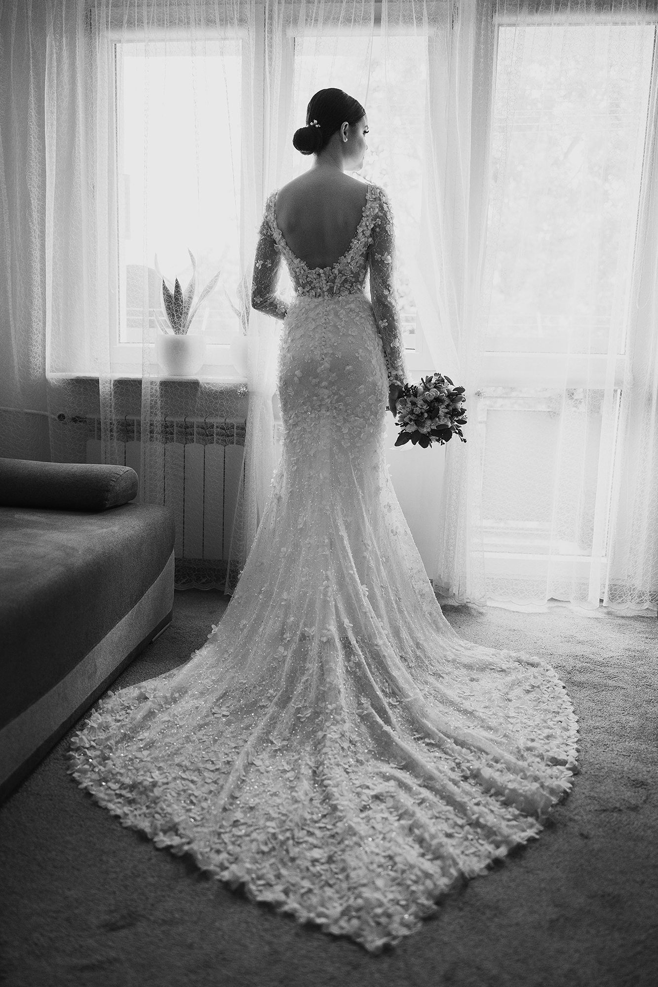 Black and white photo of bride in elegant lace backless gown with train standing by window.