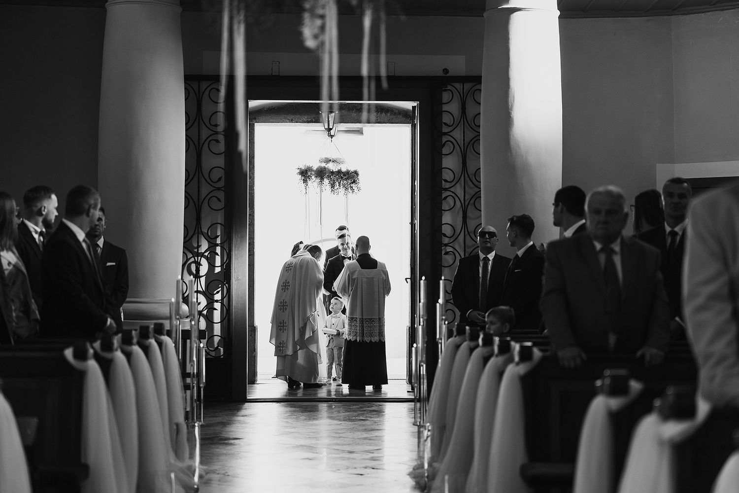 Guests line the aisle as a wedding ceremony takes place at the entrance of an elegant church in black and white.