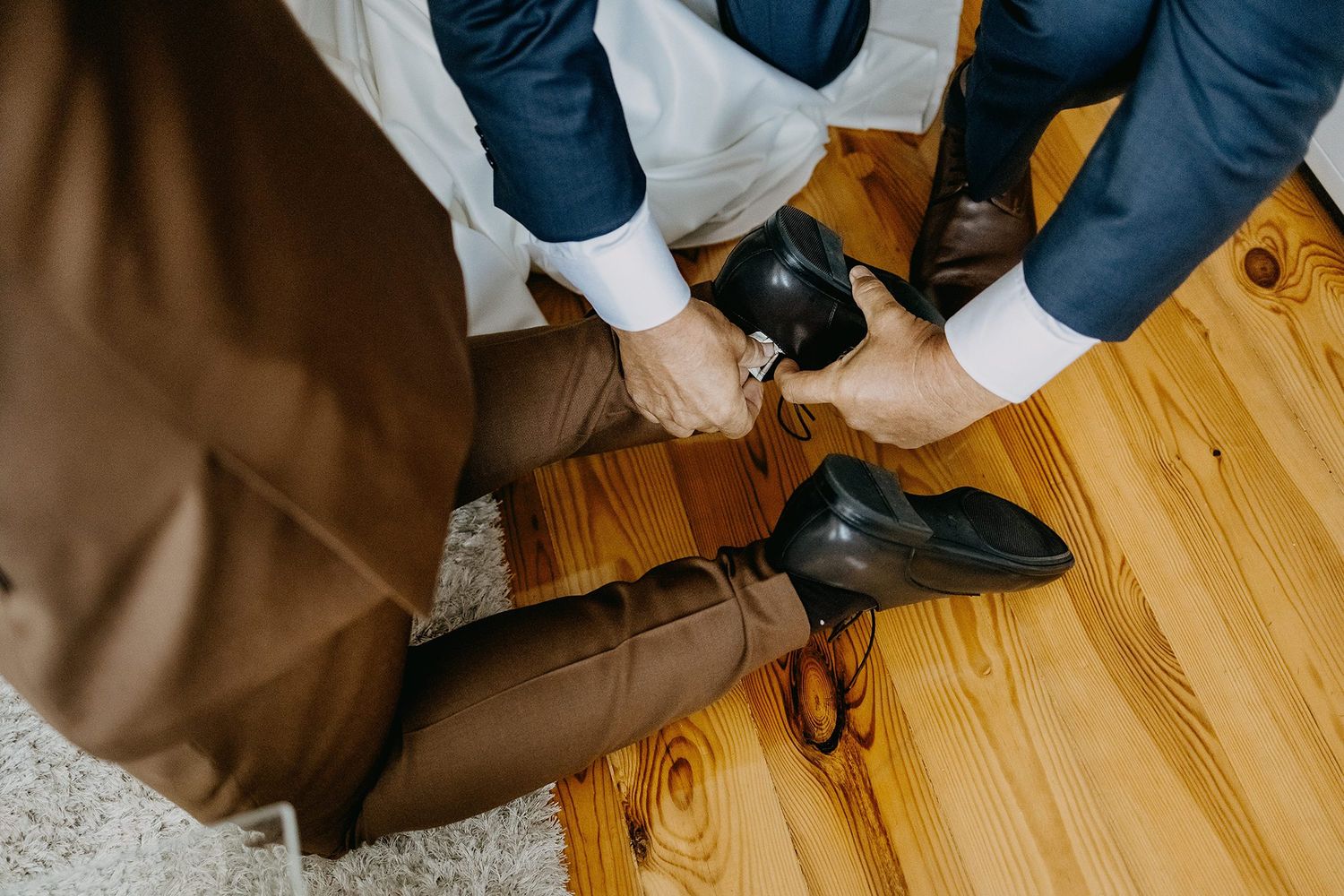 Groom being helped putting on black dress shoes on wooden floor.