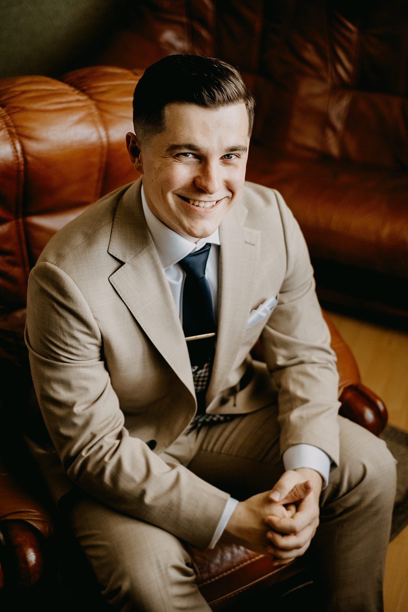 Smiling groom in beige suit and tie sitting on a leather sofa before his wedding.