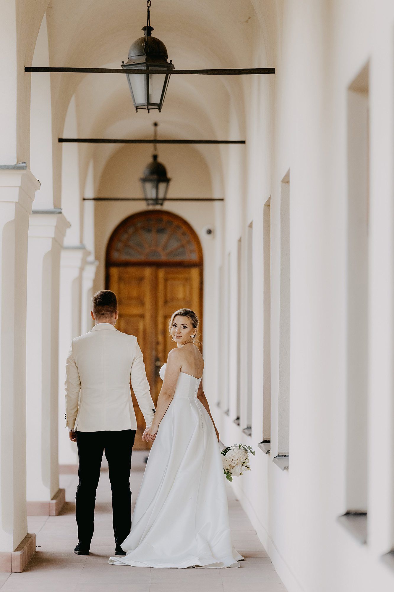 Bride glances back while holding grooms hand walking through elegant arched corridor toward ornate door.