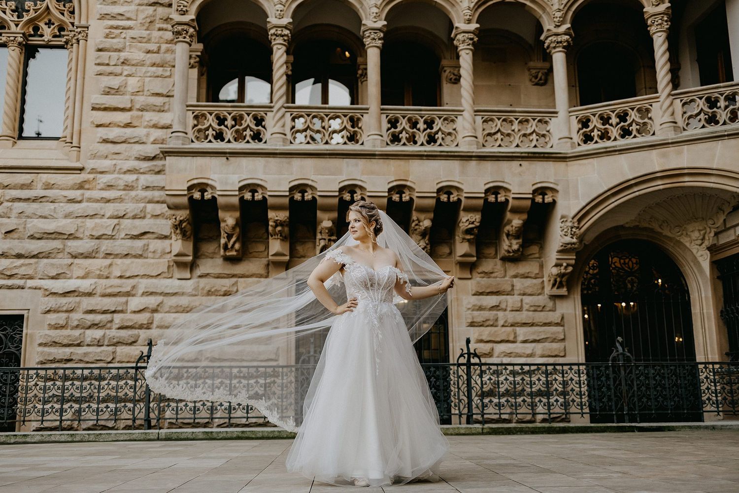 Bride in a flowing white gown with veil billowing stands in an ornate historic stone courtyard.