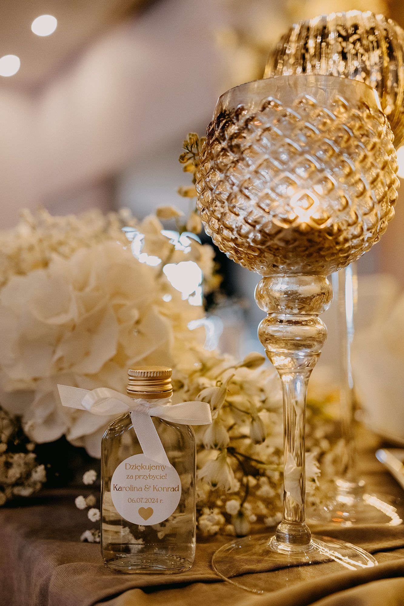 Elegant gold crystal candle holder beside a personalized wedding favor bottle with white hydrangeas.