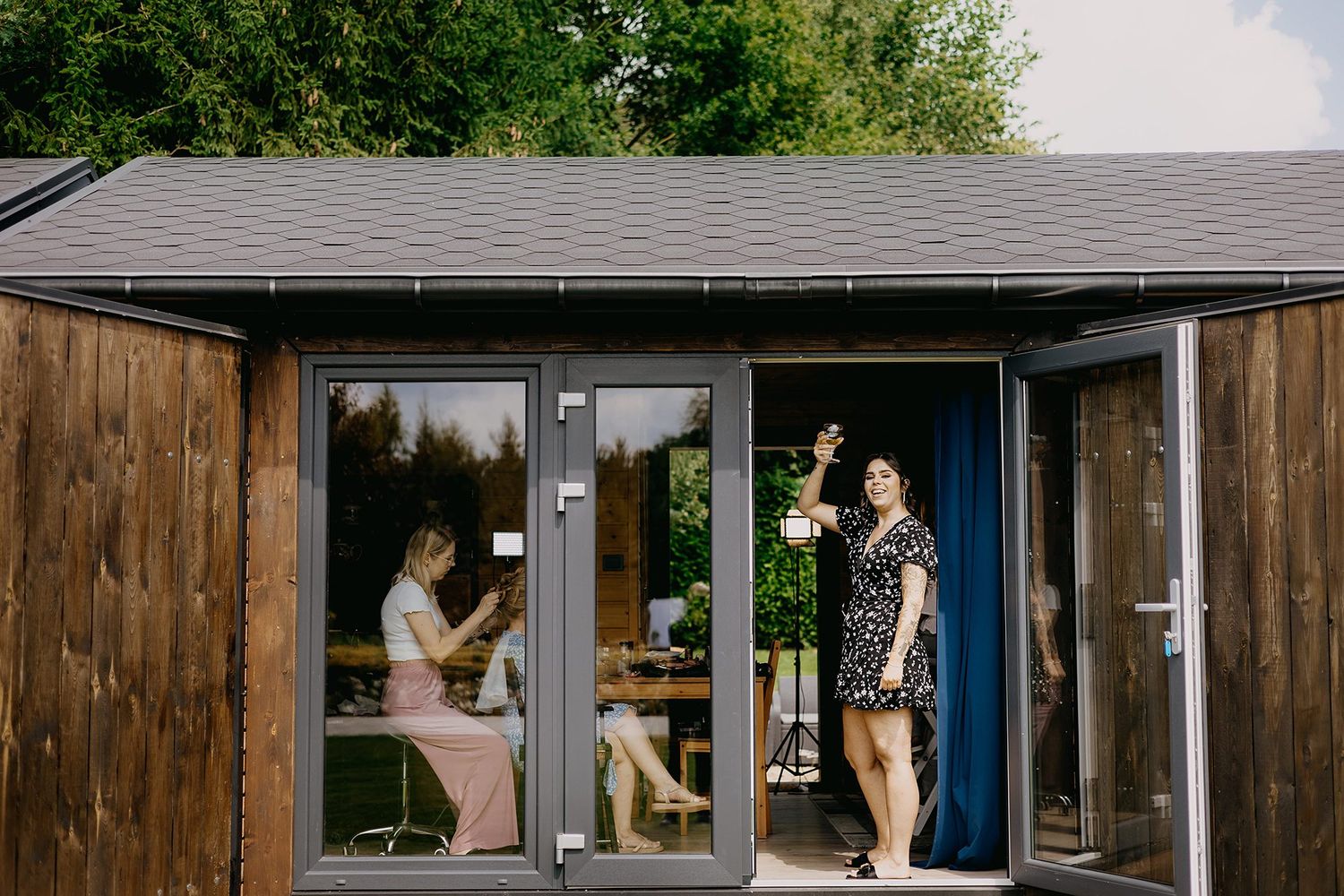 Two women relax outside modern wooden cabin with glass doors at wedding venue.