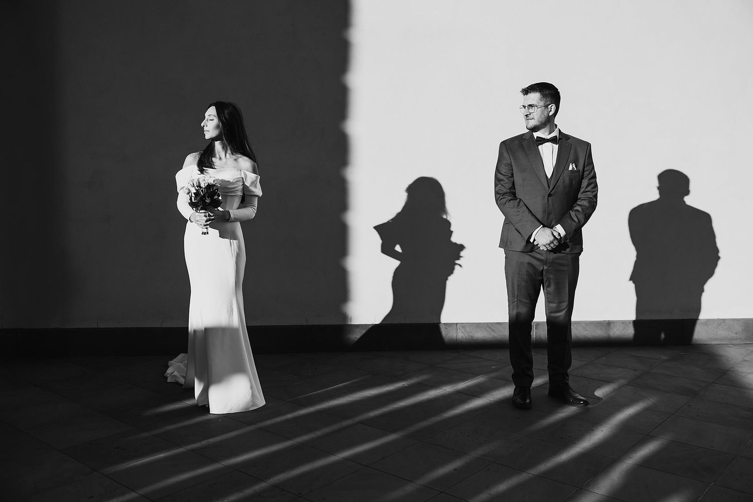 Elegant black and white wedding portrait of bride and groom standing apart with dramatic shadows on a white wall.