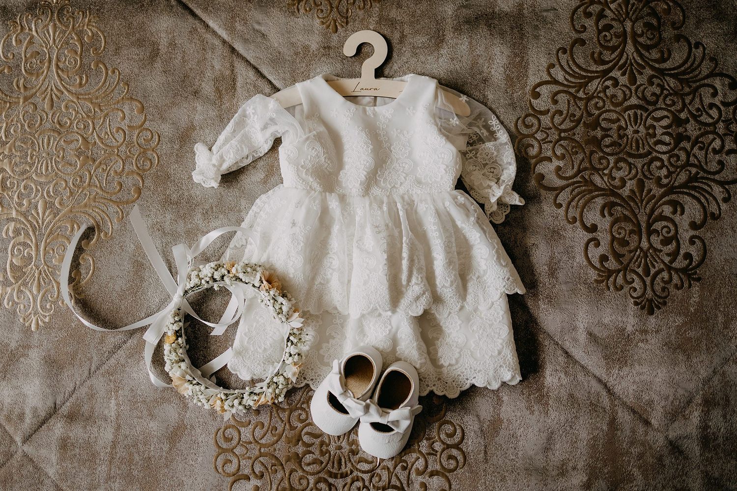 White lace baby christening dress with floral crown headband and white shoes flat lay on textured surface.