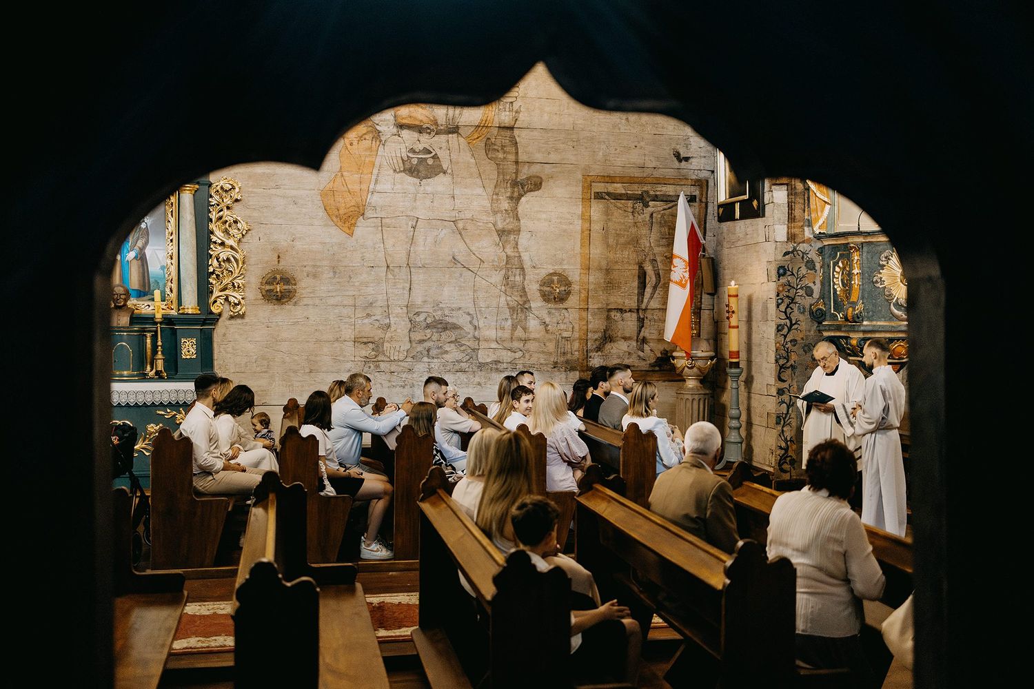 Congregation seated in wooden pews of a historic church during a baptism ceremony, viewed through an archway.