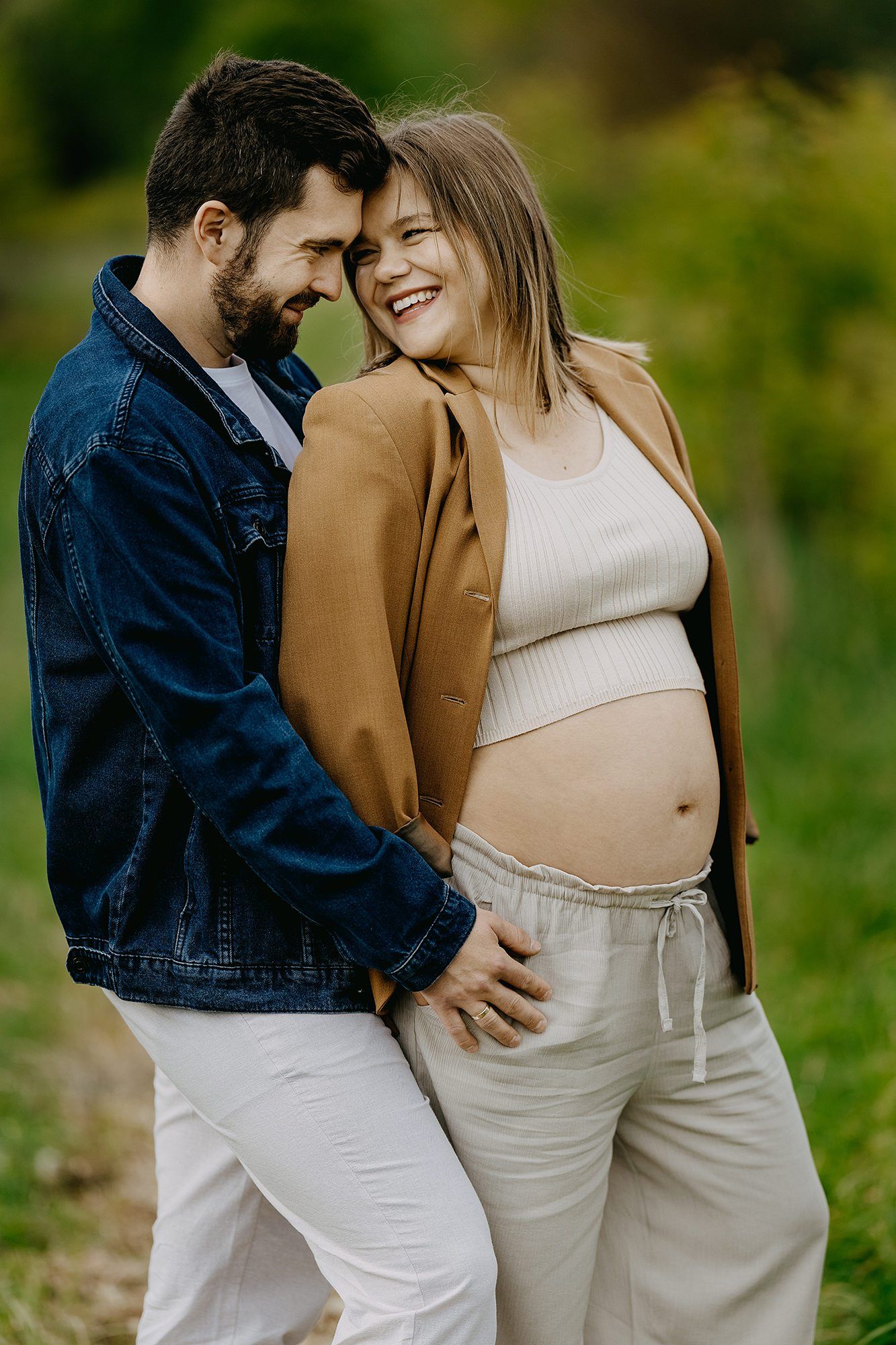Laughing pregnant couple embracing outdoors, woman showing baby bump in beige outfit with brown blazer.