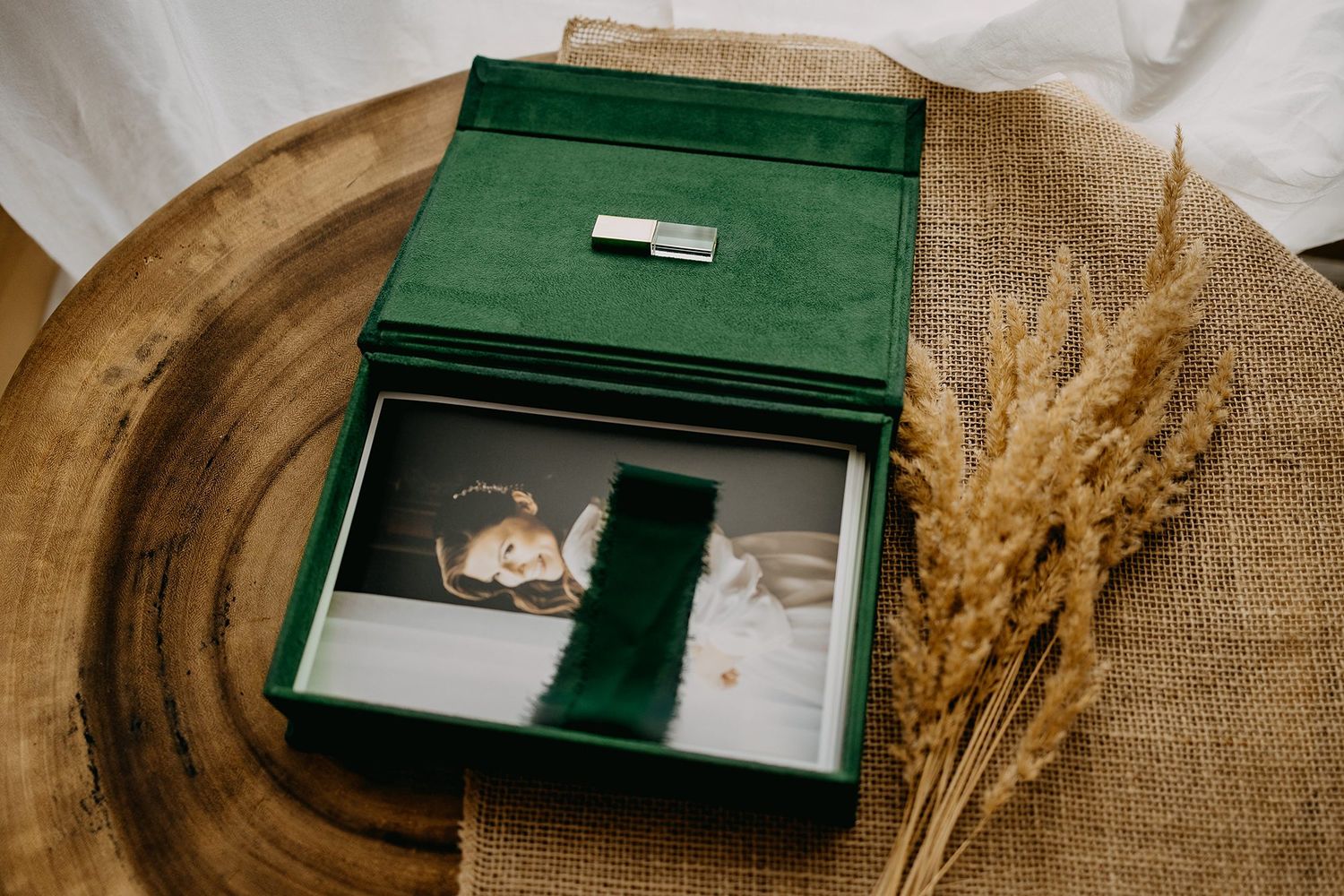 Open emerald green velvet wedding photo box with USB drive and dried wheat on wooden surface.