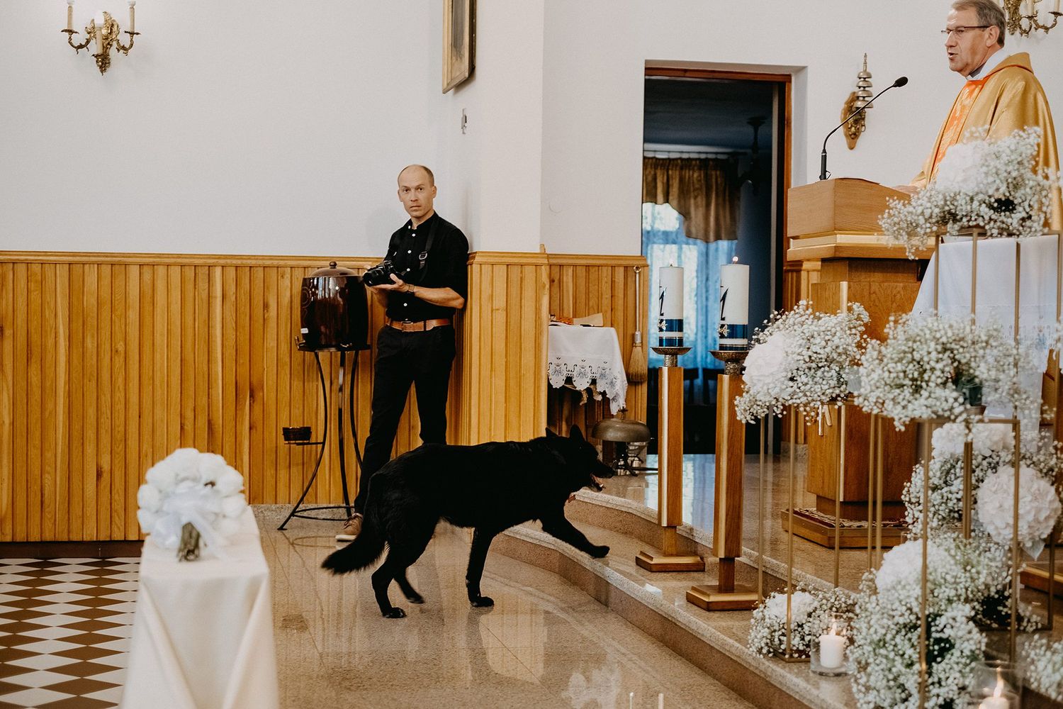 Black dog walking past a man in clergy attire inside a decorated church during a ceremony.