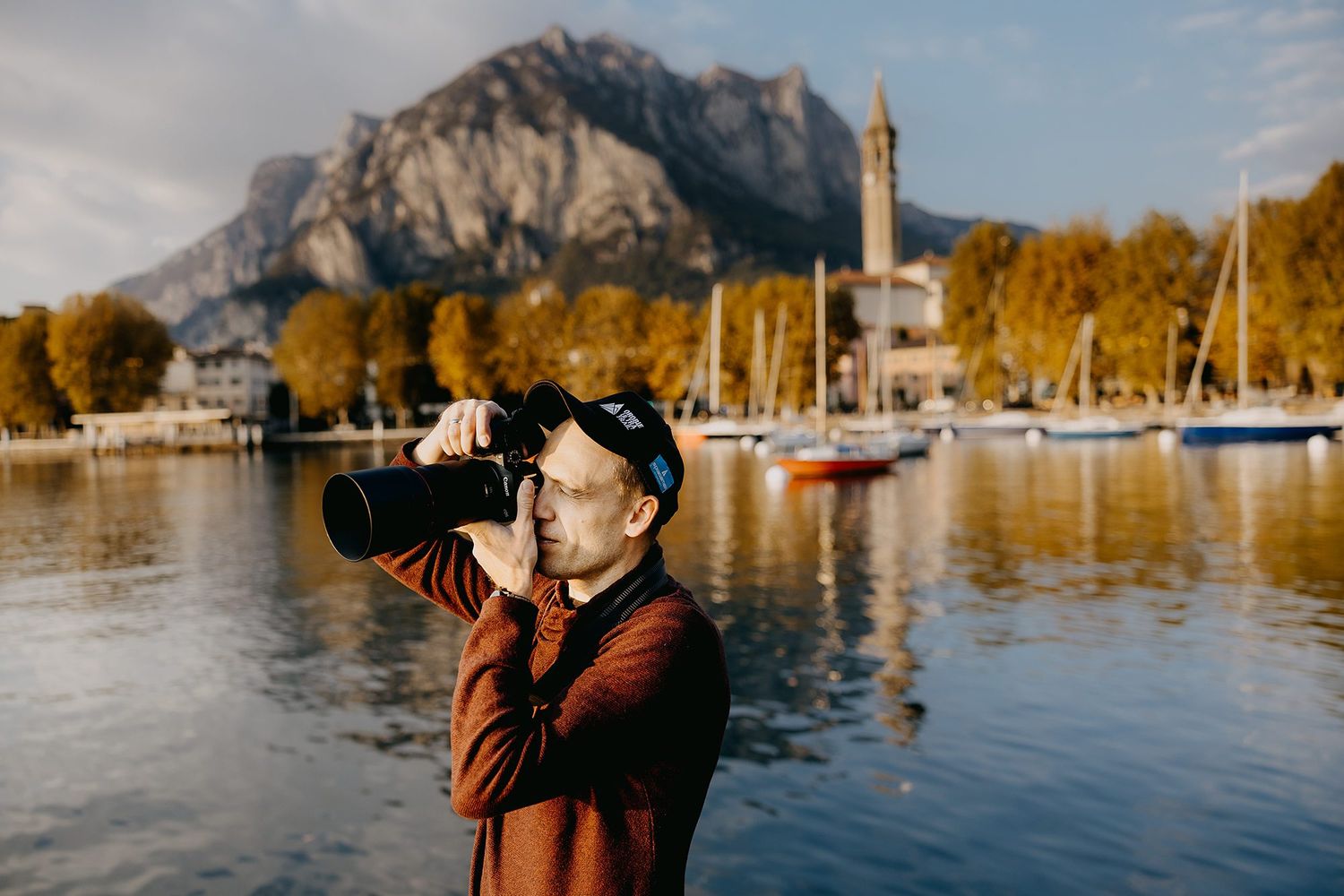 Man photographing scenic lakeside marina with mountain church backdrop during golden autumn sunset.