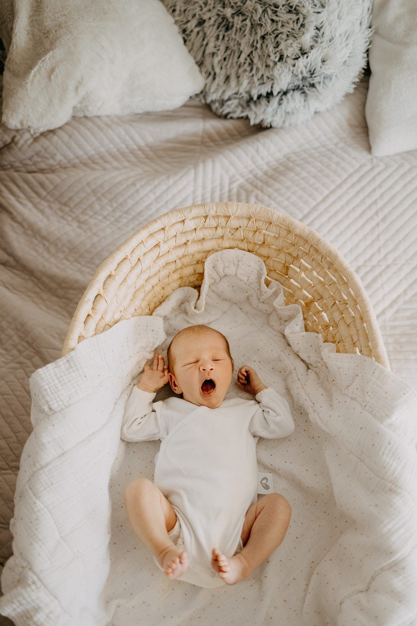 Adorable newborn baby yawning while lying in a wicker moses basket on a cozy white bed.