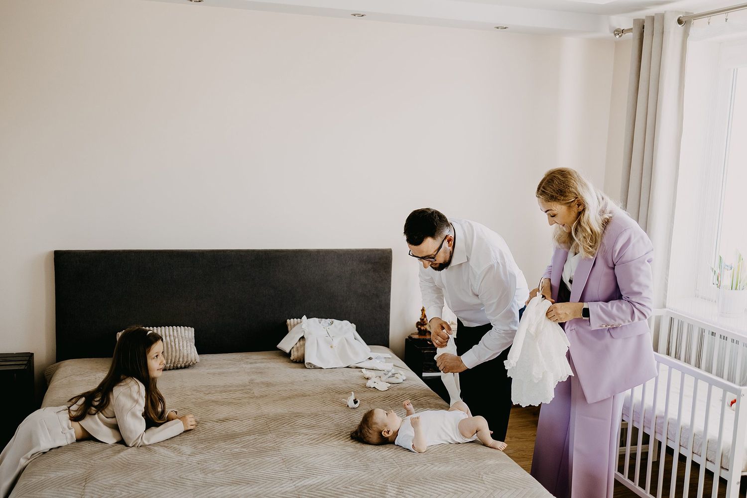 Two parents and a young girl prepare a baby in christening clothes on a bed at home.