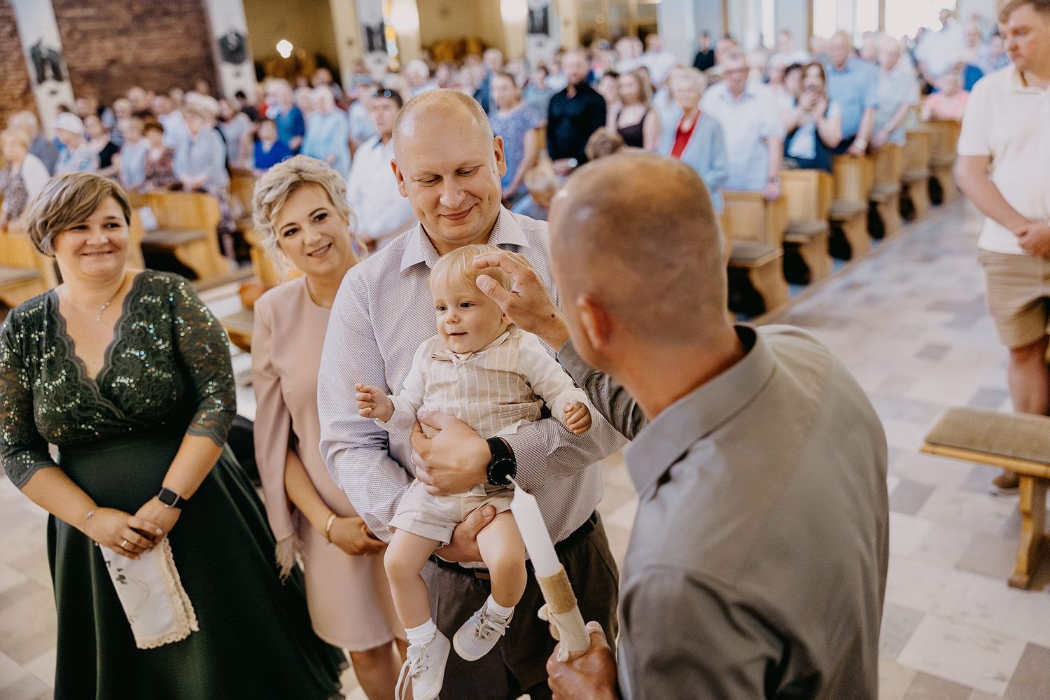 Bald grandfather proudly holding baby during christening ceremony while family smiles in church.