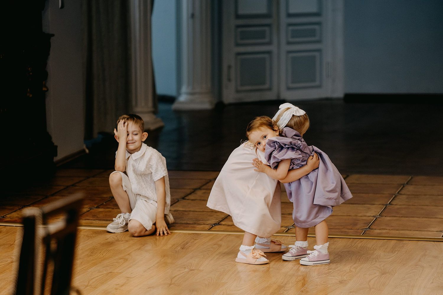 Two young girls in elegant dresses play together on a wooden floor during a christening party.