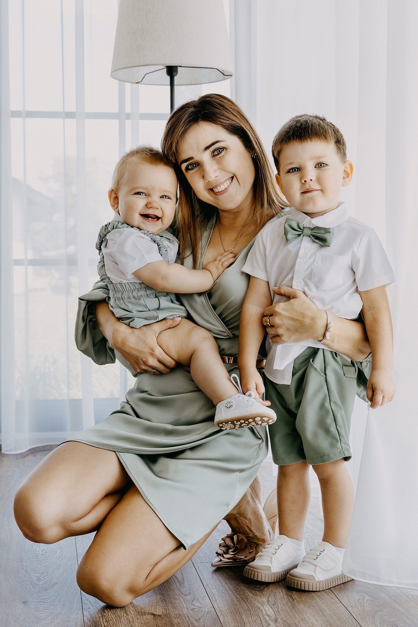 Smiling mother in sage green dress holding two young children dressed in coordinating outfits indoors.