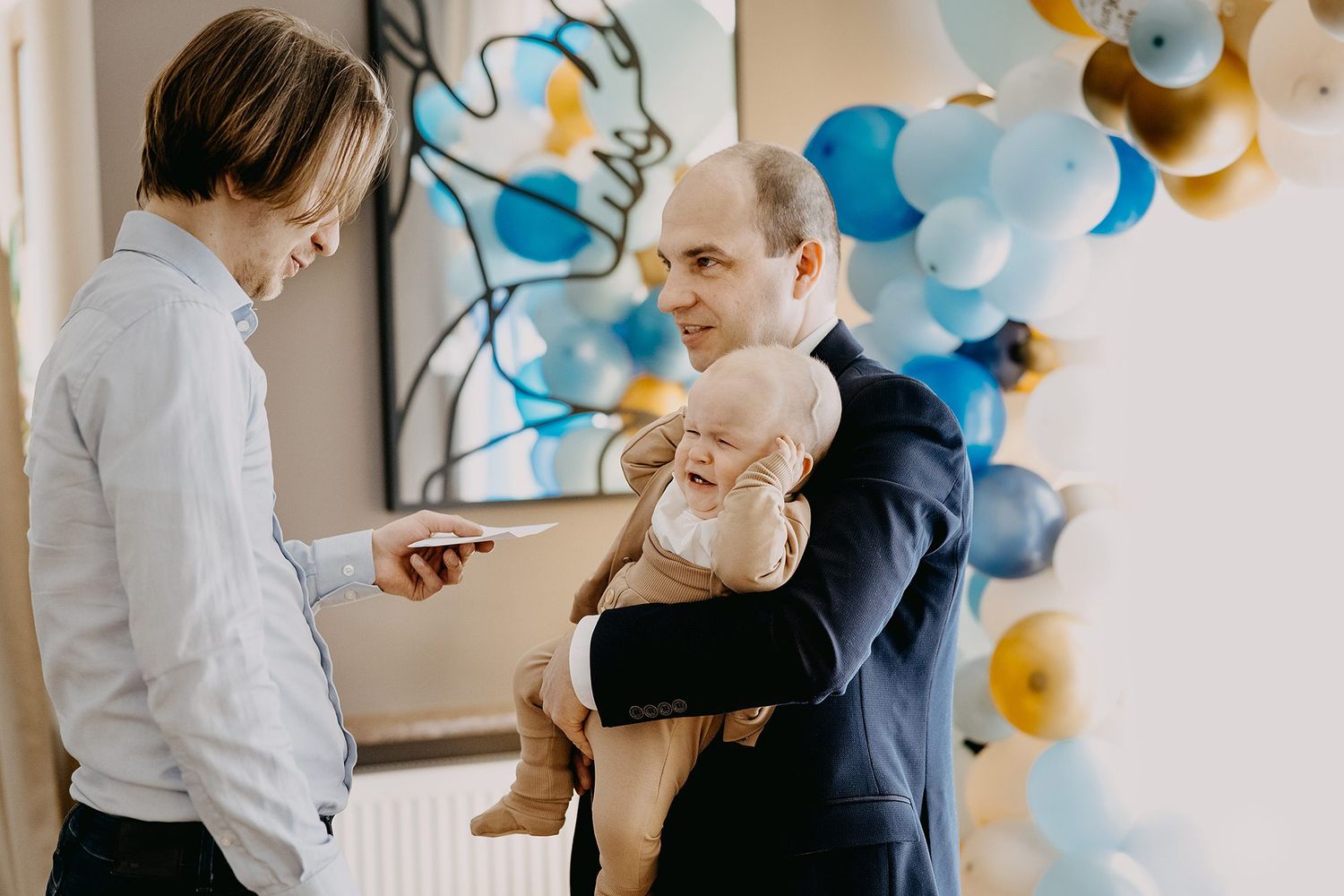 Father holding baby at christening party with blue and gold balloon arch decoration in background.
