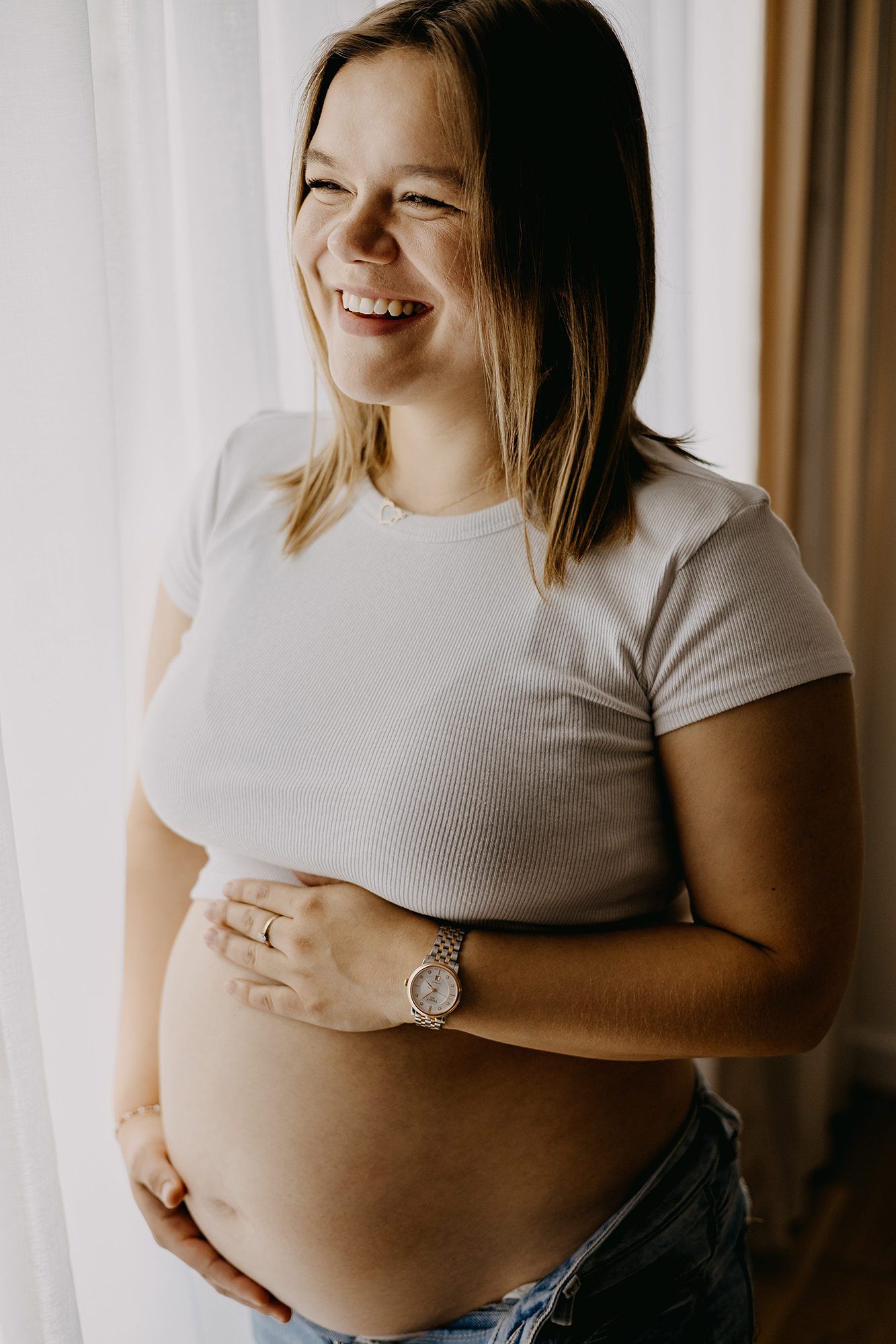 Happy pregnant woman in white t-shirt laughing joyfully while holding baby bump near a bright window.