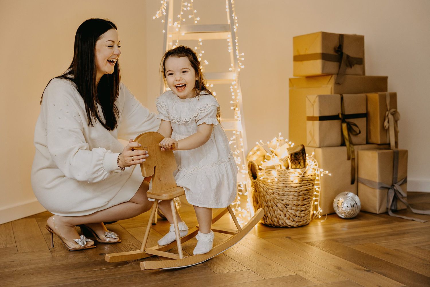 Mother and daughter laughing joyfully beside a wooden rocking horse with Christmas gifts and fairy lights in background.