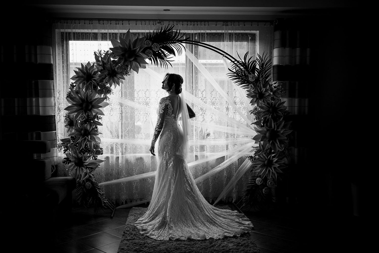 Black and white silhouette of bride in lace gown standing in circular floral arch by window.