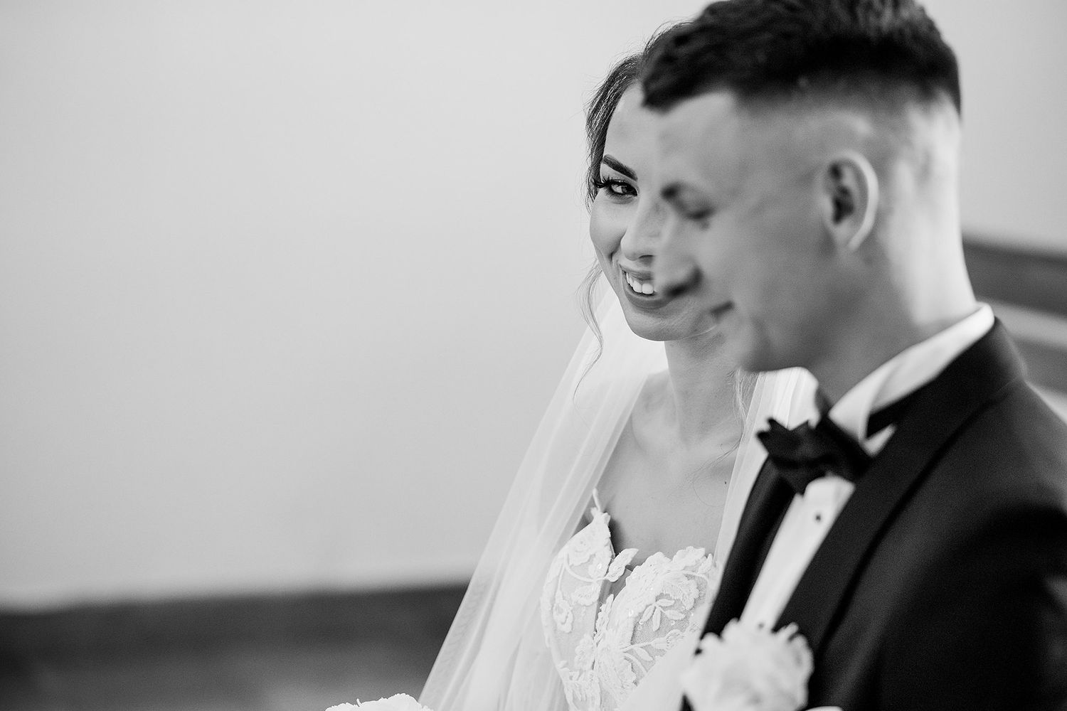 Black and white portrait of smiling bride in veil beside groom in tuxedo during wedding ceremony.
