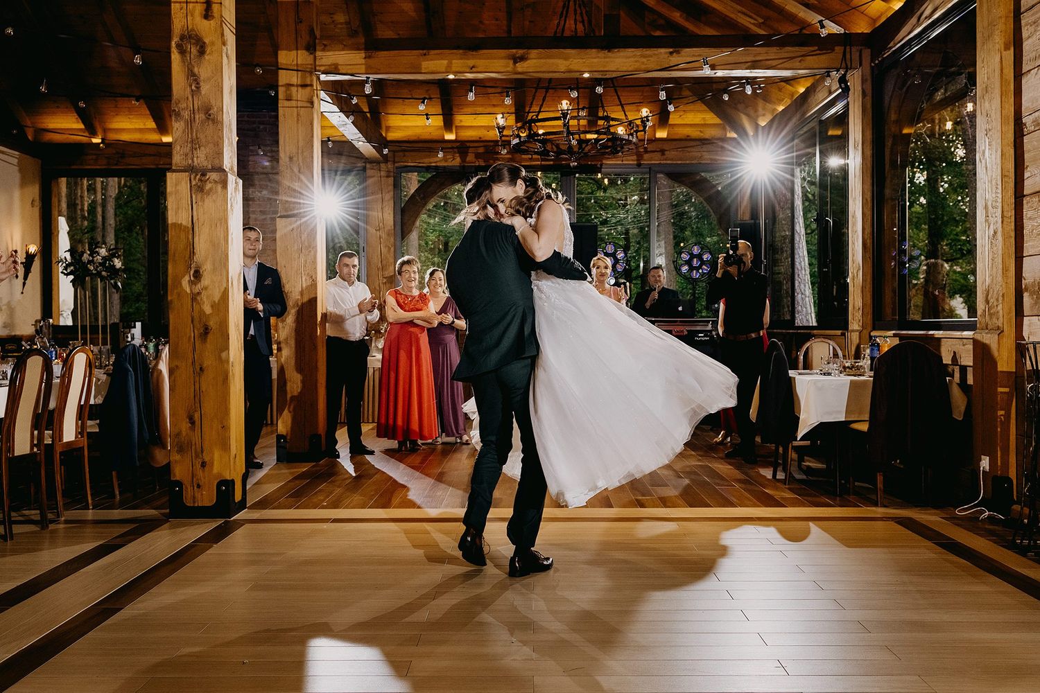 Groom twirls bride in flowing white dress during first dance in elegant wooden reception hall.