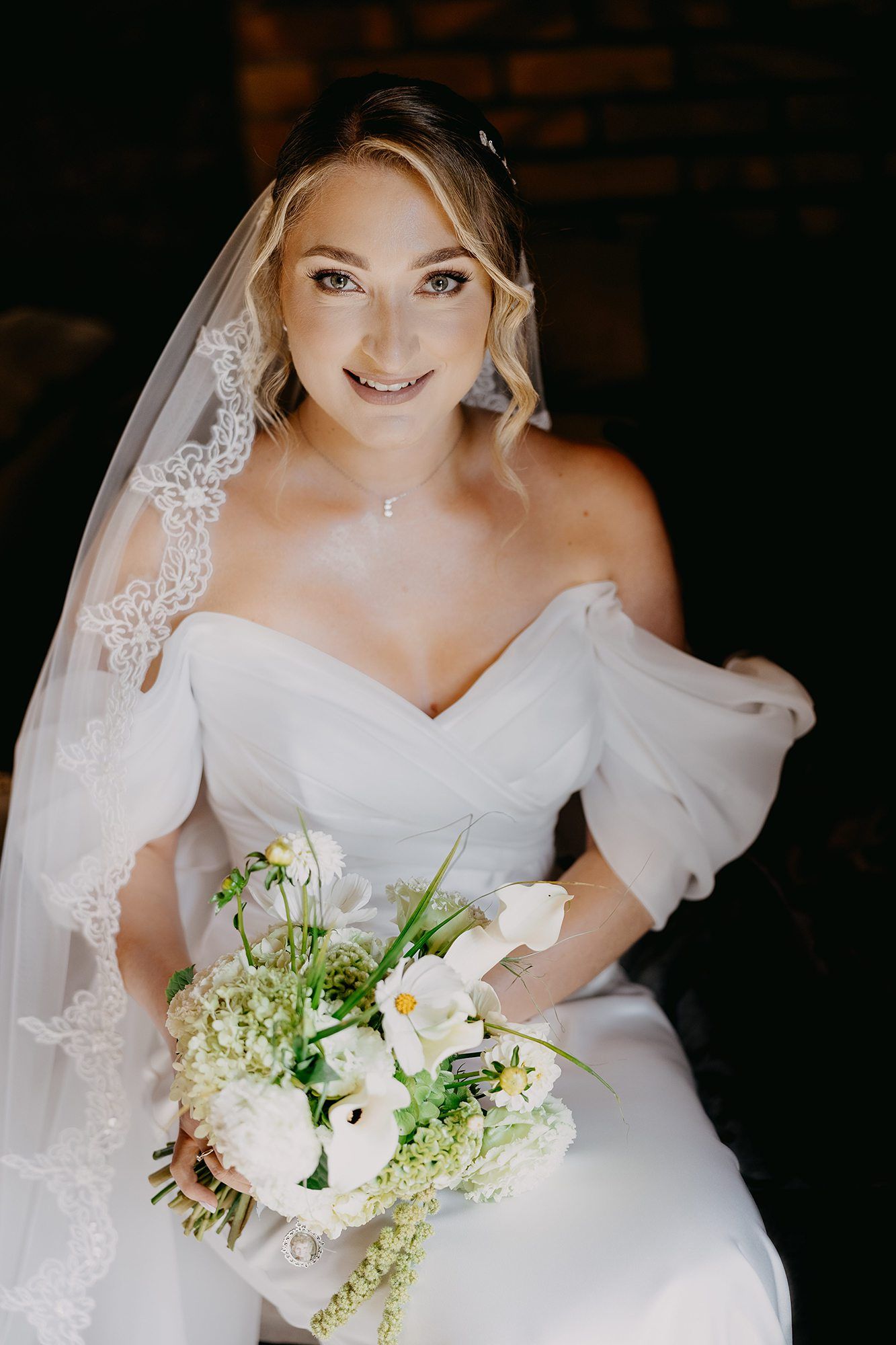 Smiling bride in off-shoulder white gown holds white floral bouquet wearing a lace-trimmed veil.