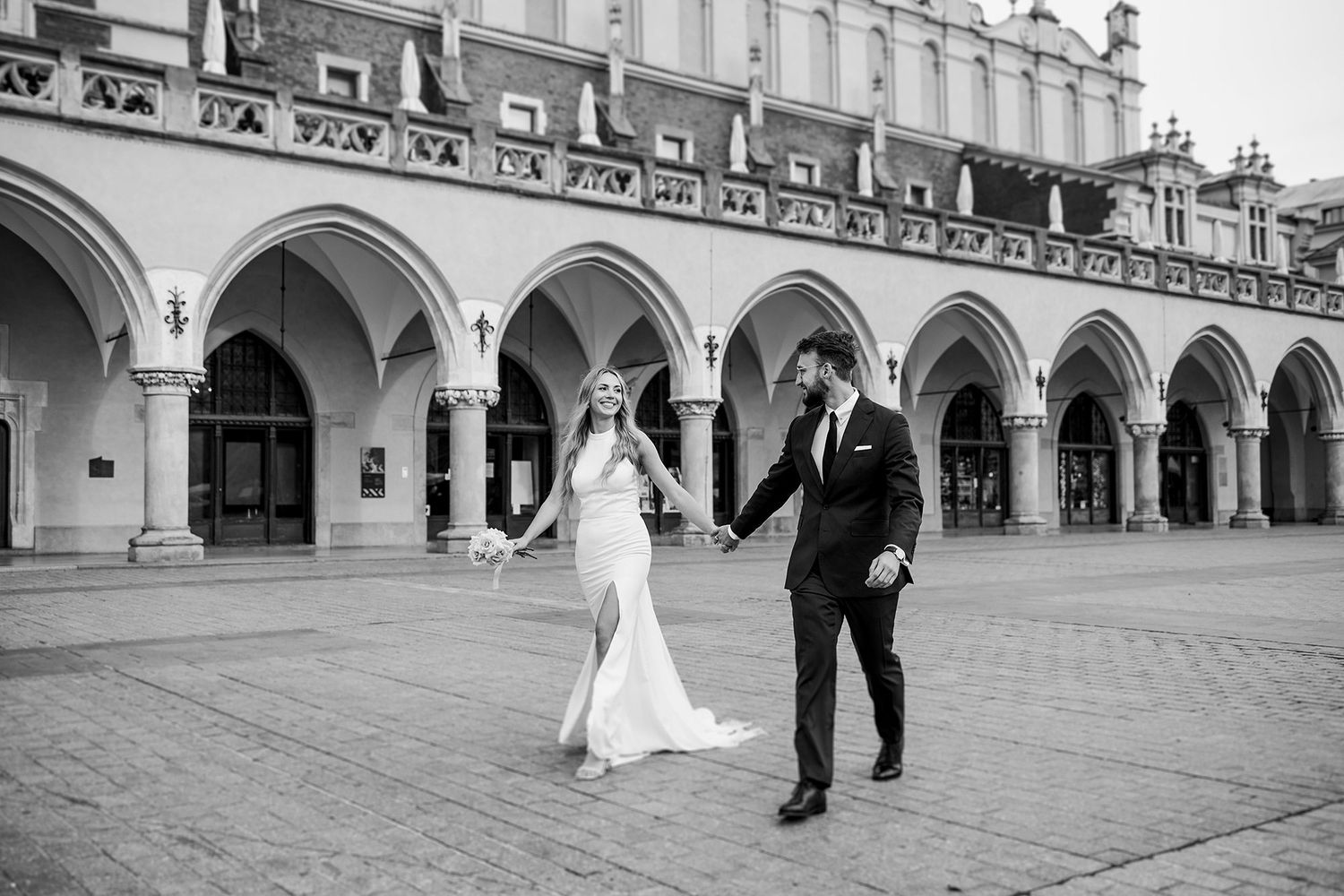 Newlyweds walking hand in hand before Gothic arches in black and white photo.