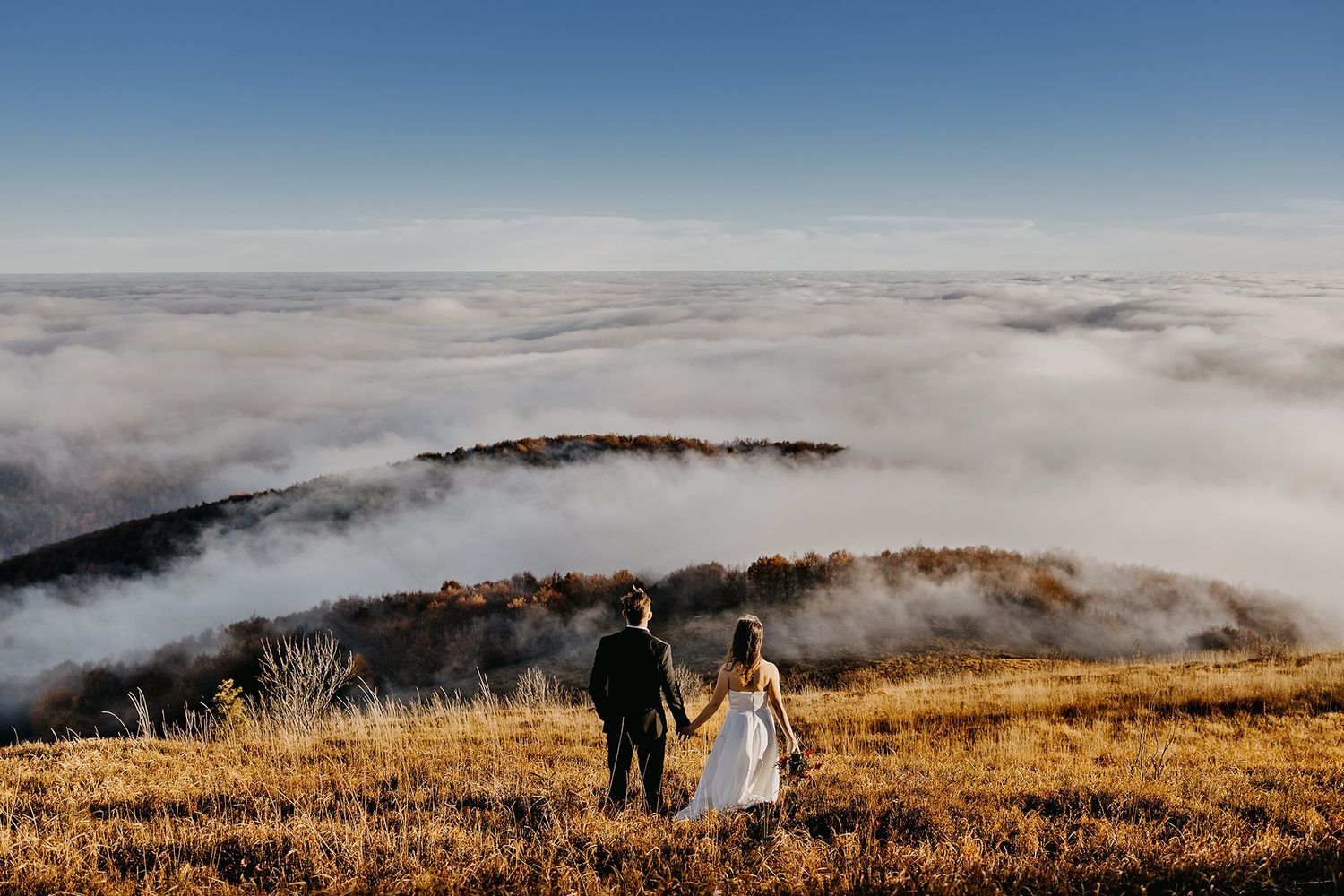 Newlyweds holding hands overlooking breathtaking sea of clouds from hilltop.