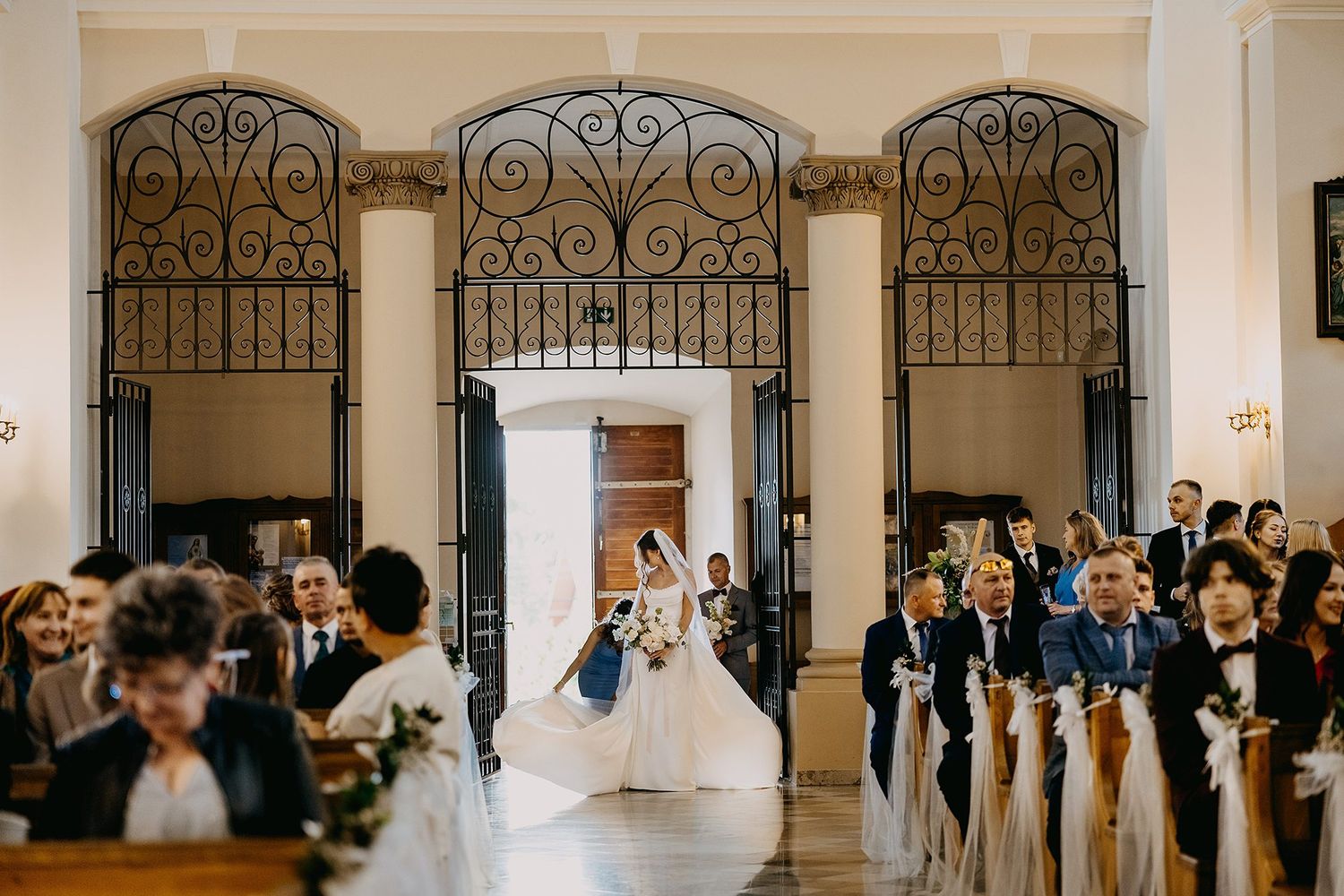 Bride walking down the aisle in a cathedral-style church with ornate iron gates and seated guests.