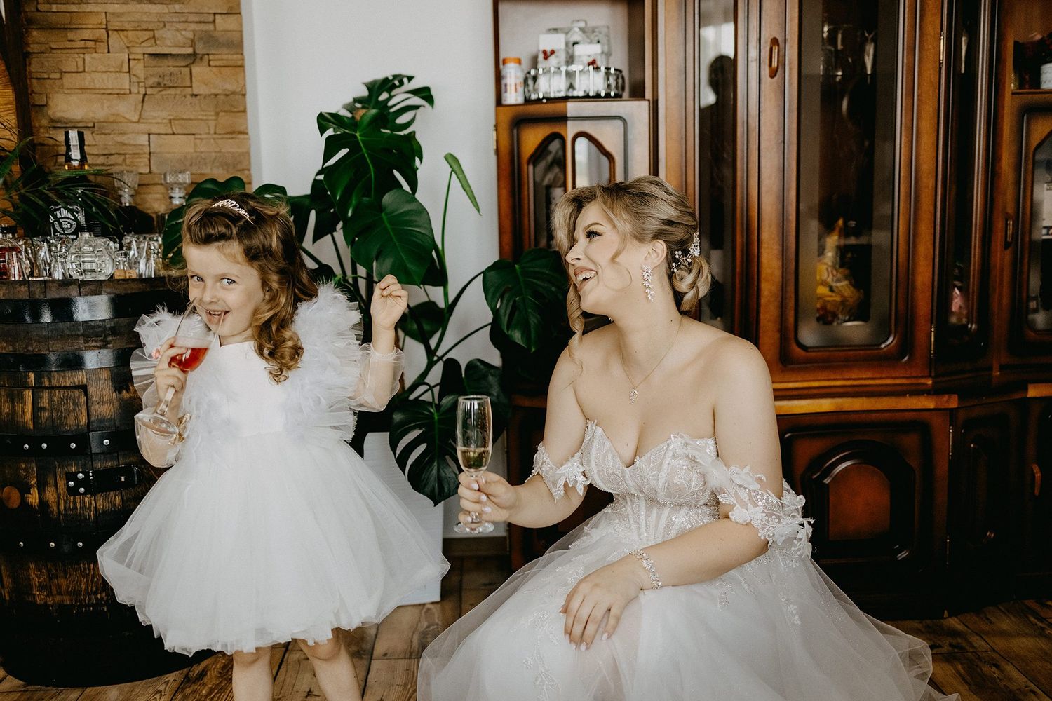 Bride in white gown smiling at flower girl in white dress holding champagne flute.