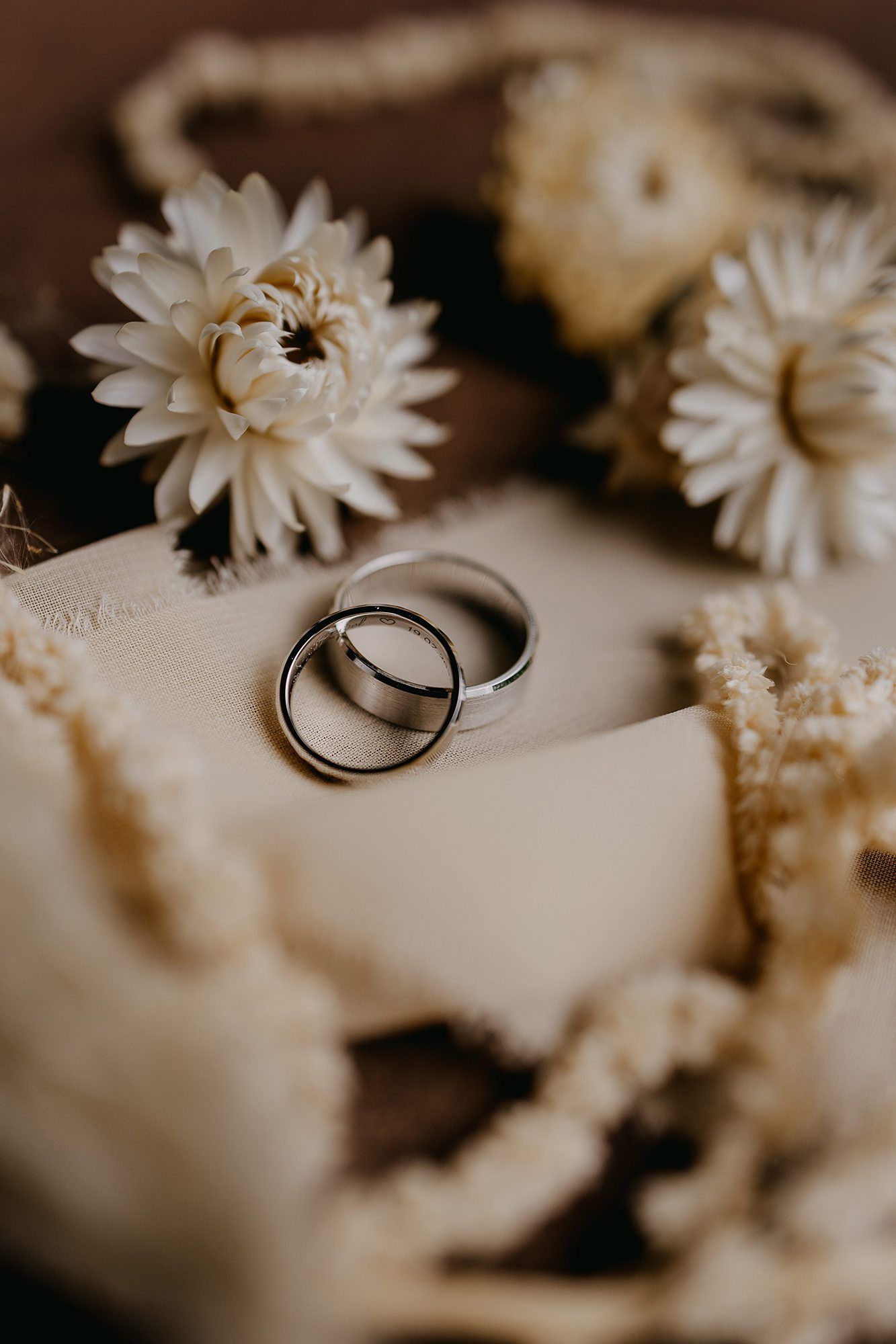Two silver wedding bands rest on a cream surface surrounded by white dried flowers and delicate blooms.