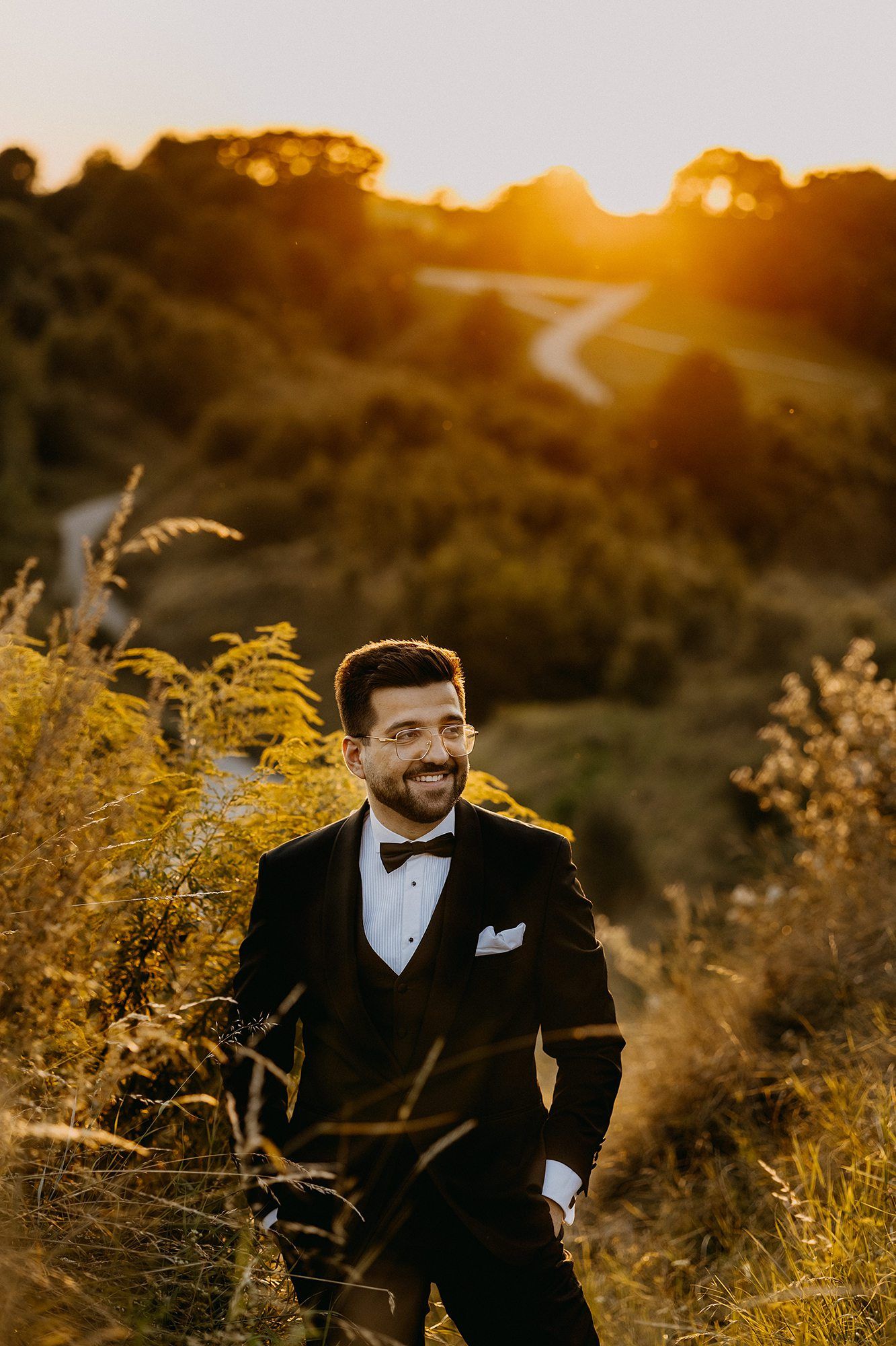 Smiling groom in black tuxedo stands in golden meadow grass at sunset.
