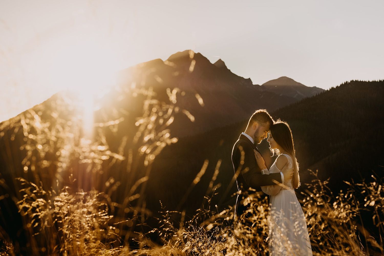 Wedding couple shares intimate kiss in golden field with dramatic mountain sunset backdrop.