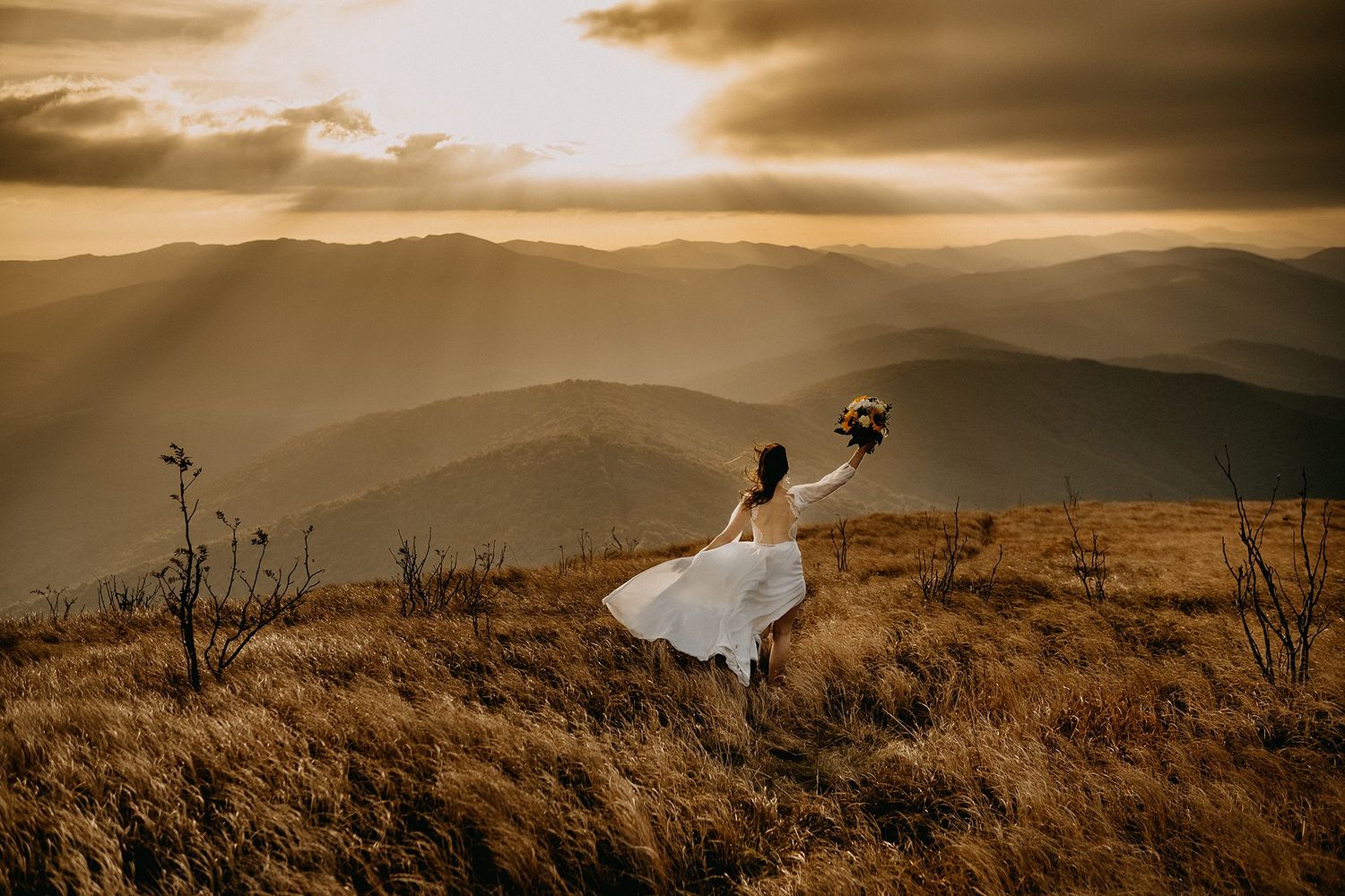 Bride in flowing white dress holding bouquet aloft on golden mountain hillside at dramatic sunset.