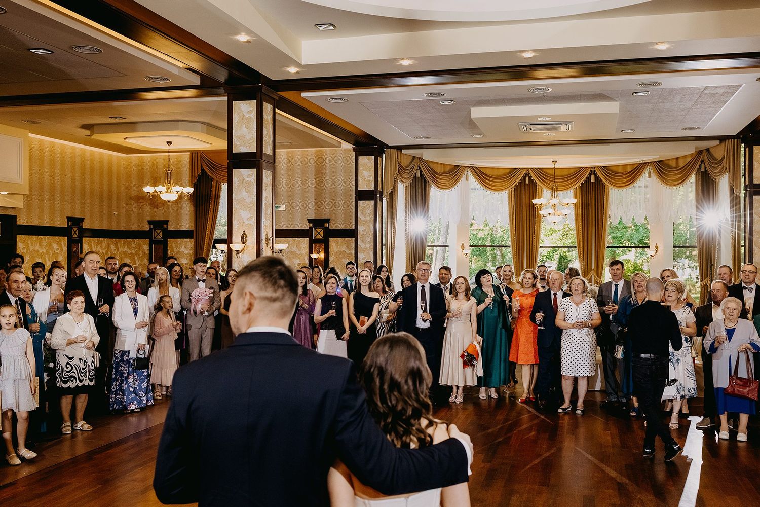Bride and groom facing wedding guests gathered in elegant ballroom during reception celebration.