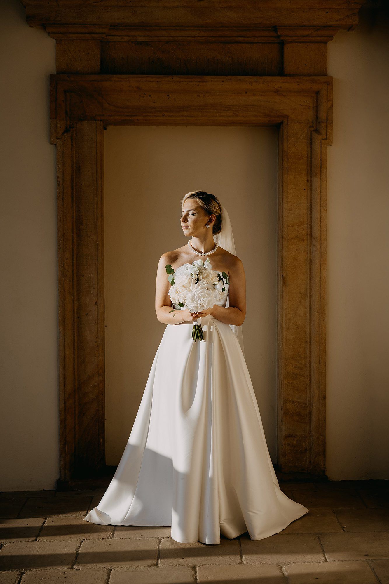 Beautiful bride in a white halter gown holding a bouquet, standing in a wooden doorframe.