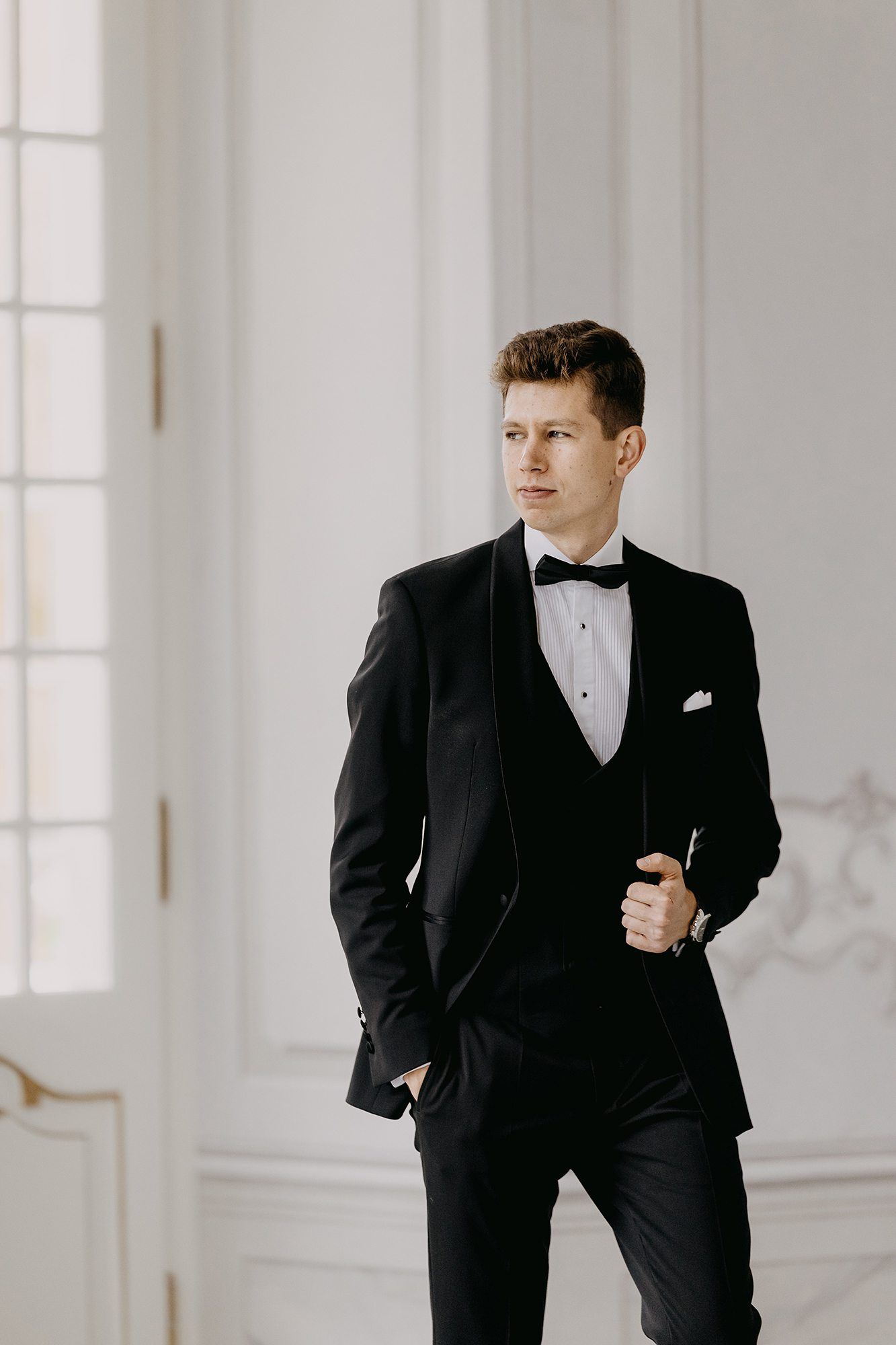 Elegant groom in black tuxedo with bow tie posing confidently in a bright white classic interior.