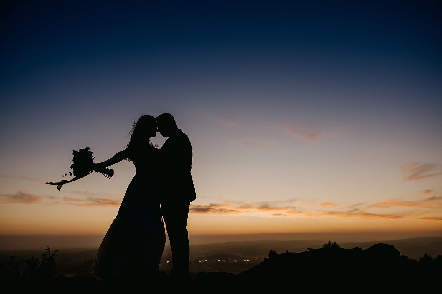 Silhouette of bride and groom sharing a romantic moment at sunset on a hilltop.