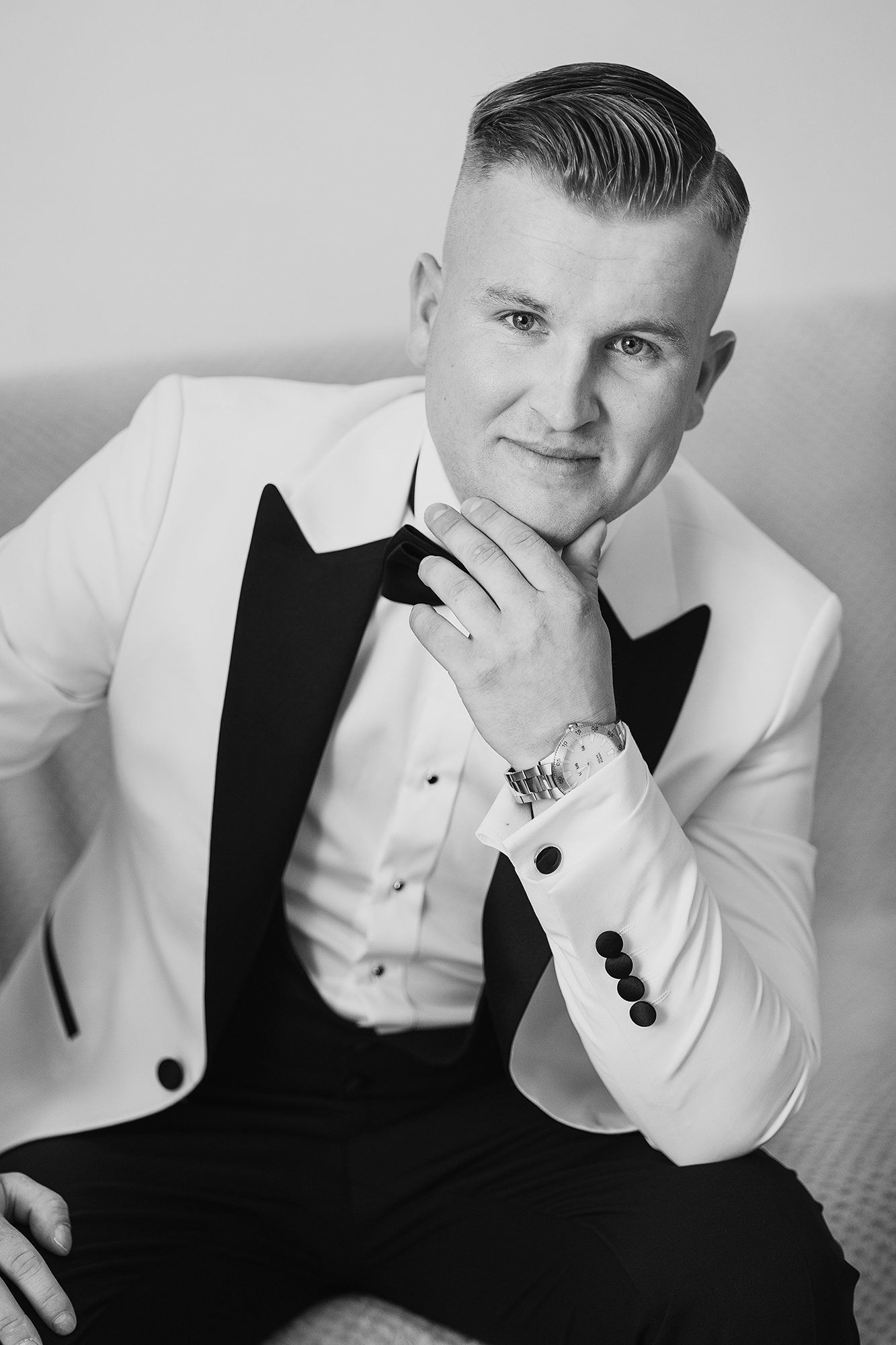 Elegant groom in white tuxedo poses confidently in black and white portrait photo.